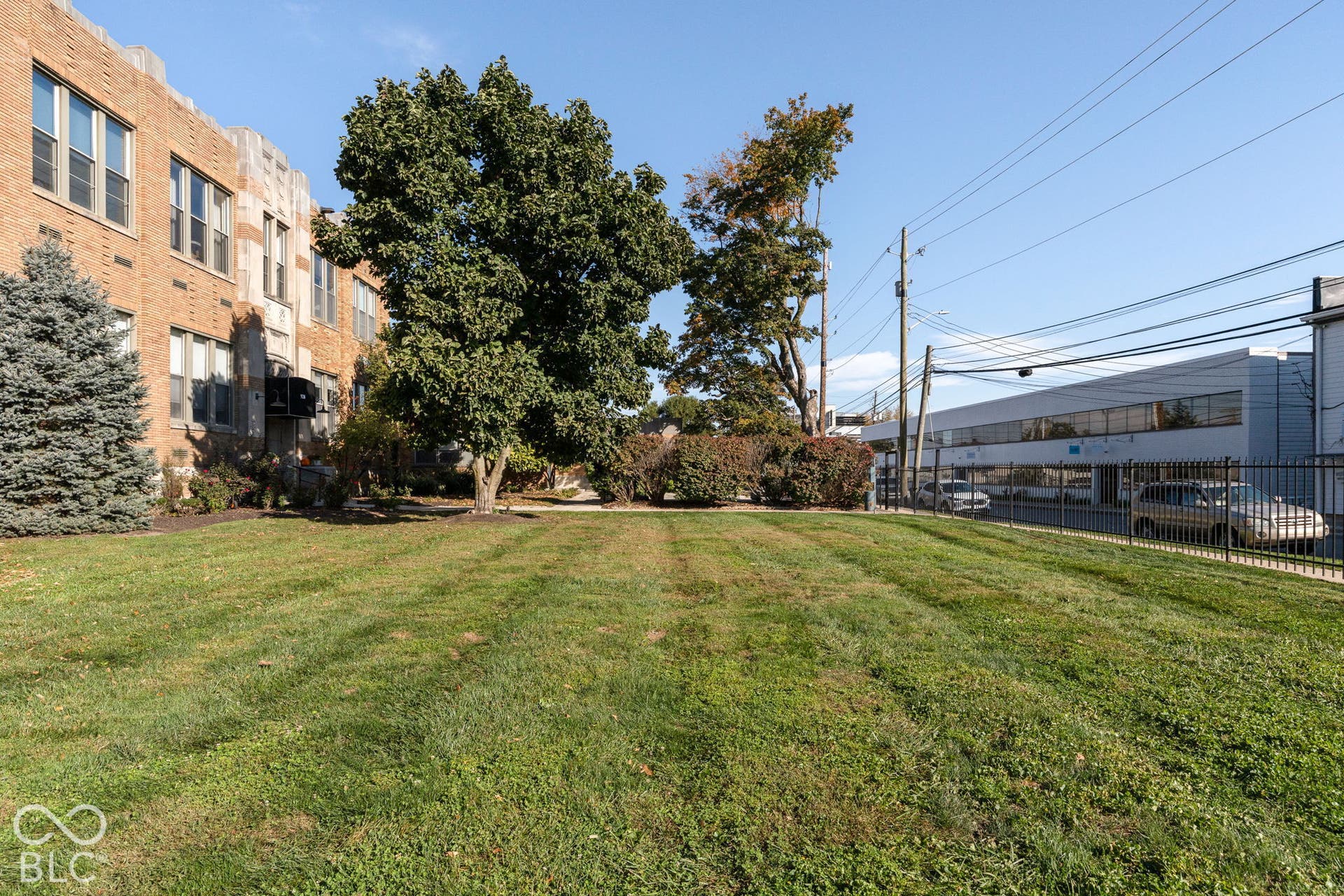 Open lot featuring a flat grassy expanse, mature trees, and a masonry exterior building facade
