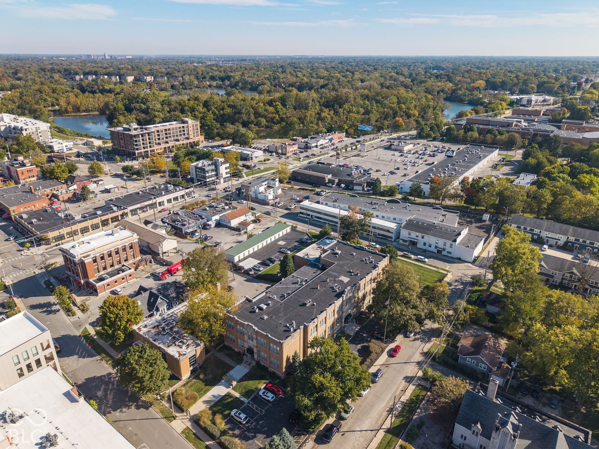 Aerial perspective of a multi-story brick commercial building featuring a flat roof system, asphalt parking lot, and proximity to regional urban infrastructure and wooded areas