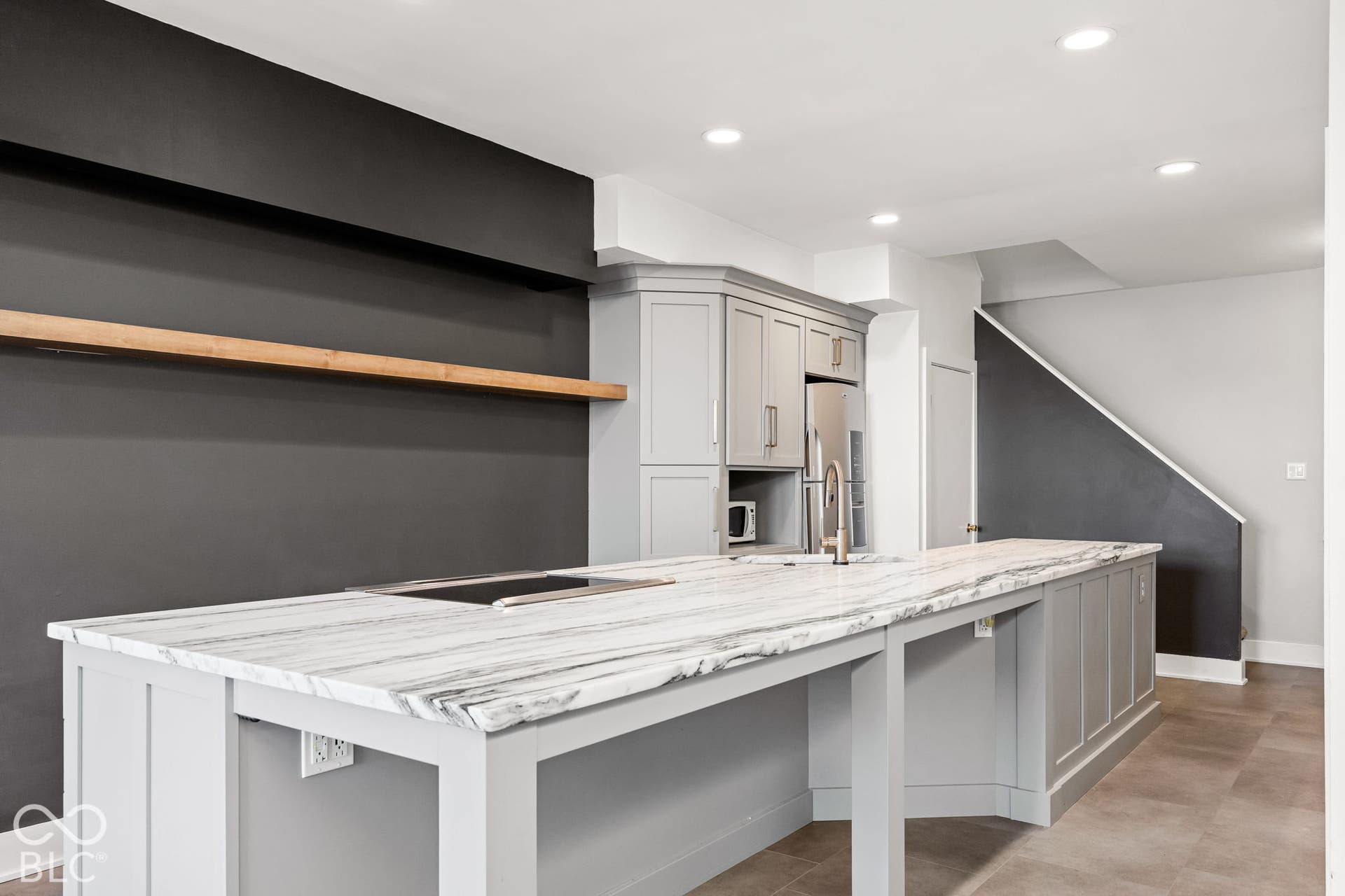 Large kitchen island with a stone countertop, grey shaker-style cabinetry, a floating wood shelf, and recessed ceiling lighting