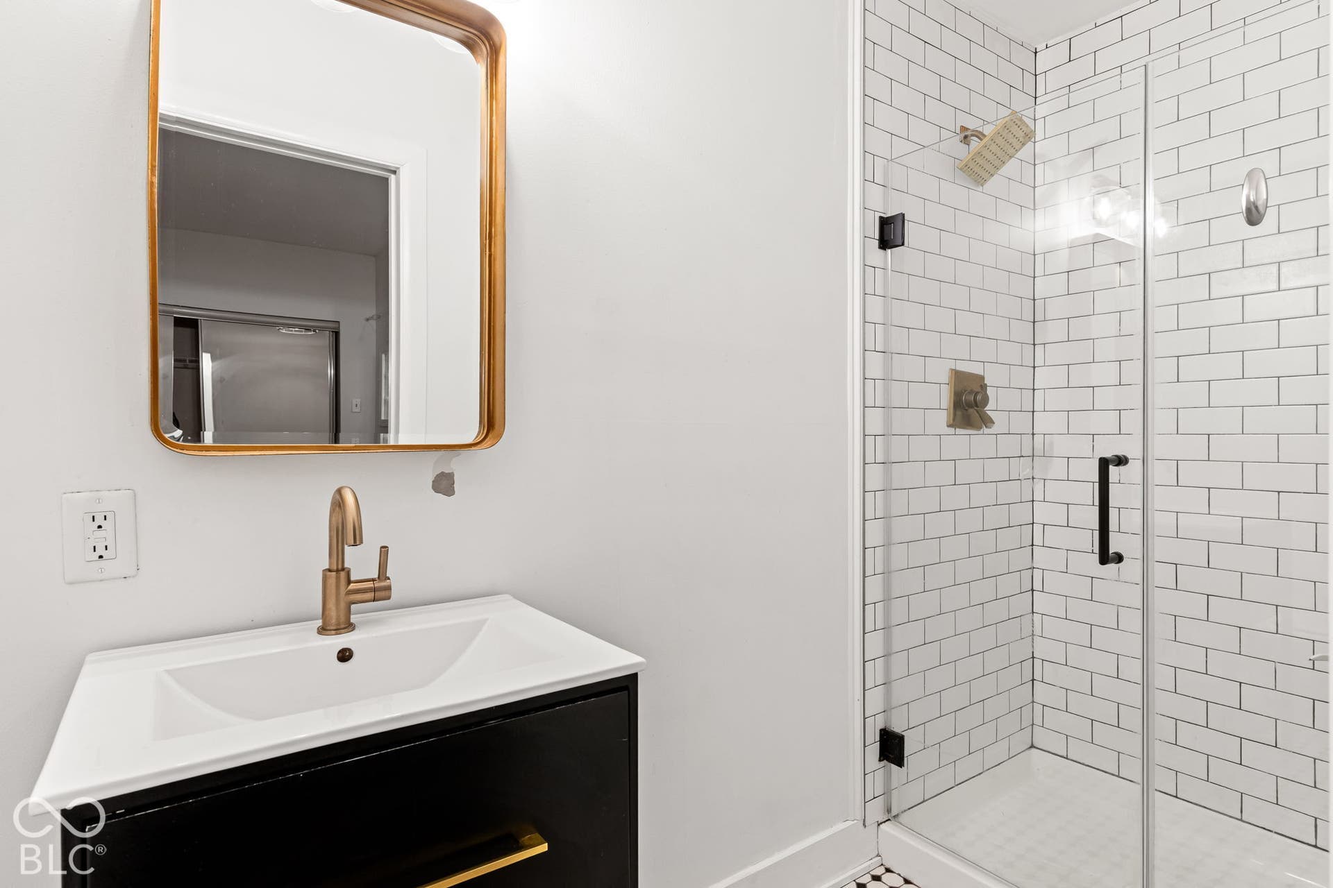 Black vanity with white countertop, gold-tone faucet, rectangular mirror with gold-tone frame, glass-enclosed shower featuring white subway tile, and matte black hardware
