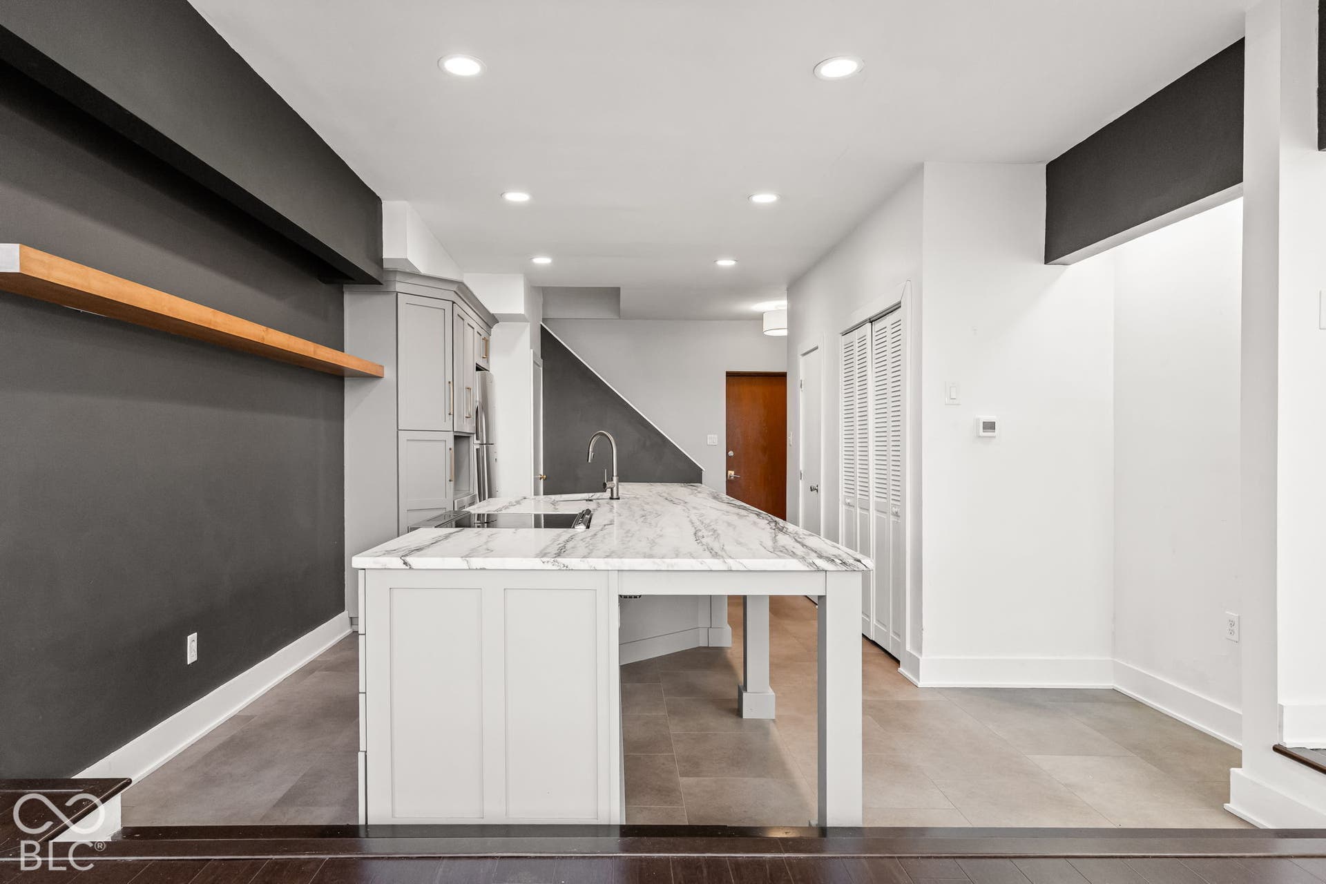 Kitchen featuring a central island with a stone-finish countertop, undermount sink, recessed ceiling lighting, wood-finish floating shelf, and neutral-tone tile flooring