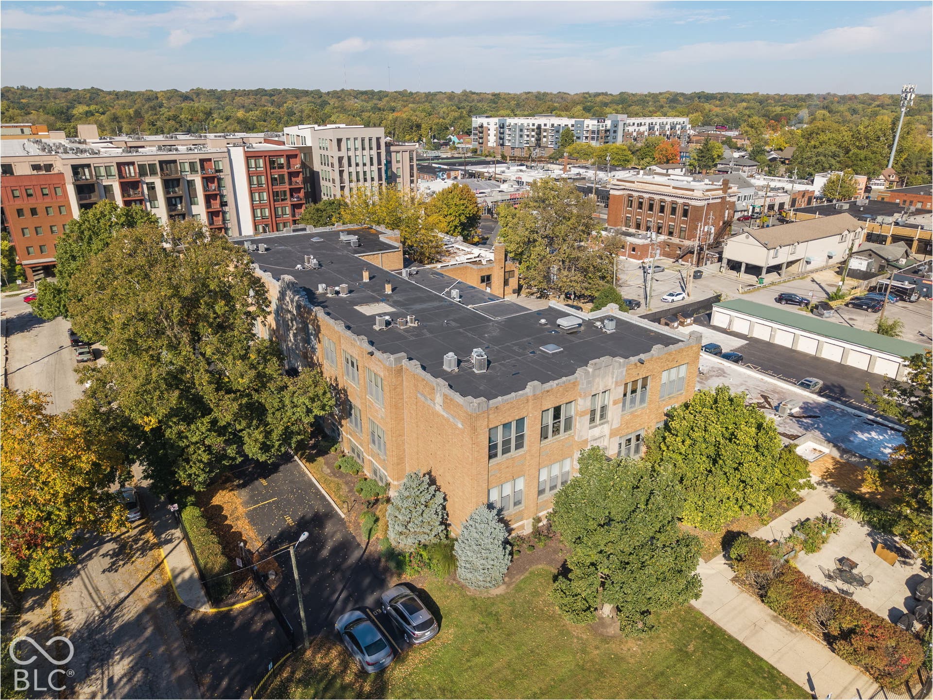 Multi-story masonry building featuring a flat roof with parapet detailing, large rectangular windows, an asphalt parking lot, and mature landscaping