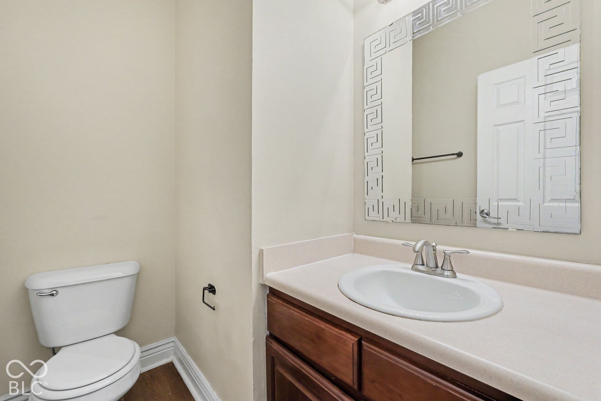 Vanity featuring a dark wood-finish cabinet, solid-surface countertop with an integrated oval basin, polished chrome double-handle faucet, and a rectangular mirror with a decorative Greek key border