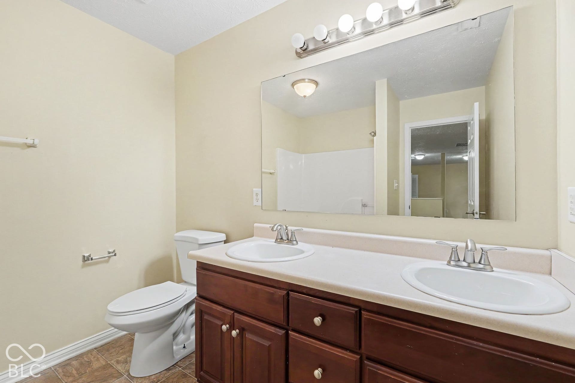 Double vanity with integrated sinks, neutral solid-surface countertop, dark wood-finish cabinetry, full-width wall mirror, and five-light vanity fixture