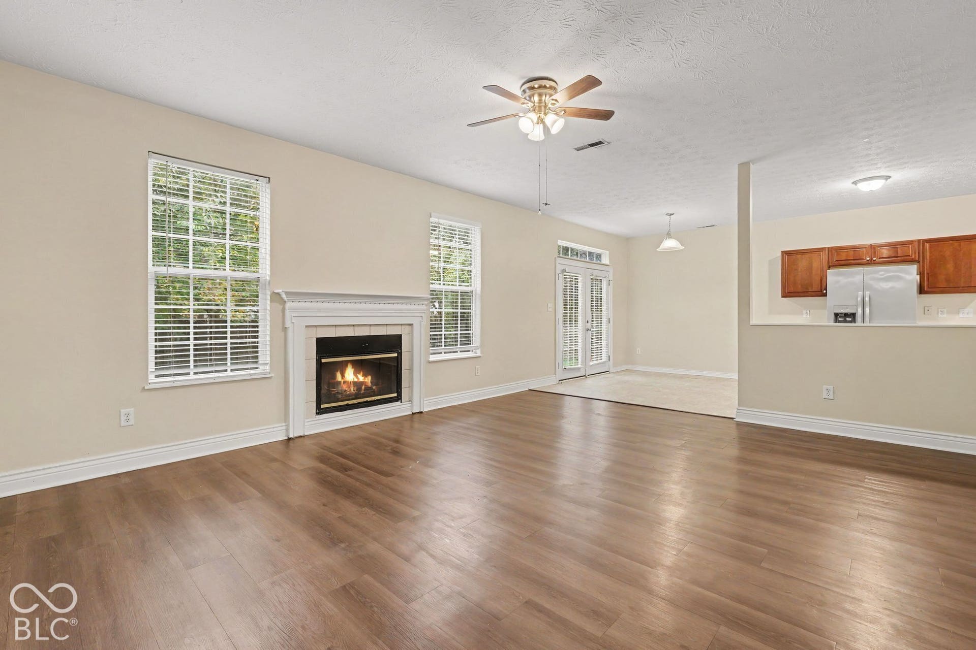 Living area featuring wood-finish flooring, a fireplace with a white mantel, ceiling fan, natural lighting through multiple windows, and glass-paneled double doors