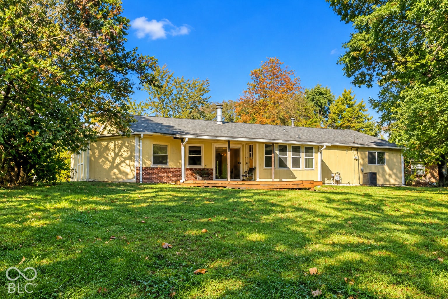 1802 Heather Court, Indianapolis, IN 46229 Virtually Altered - Exterior facade featuring light-toned siding, a partial brick veneer, a wood deck, and mature trees on a grass-covered lot