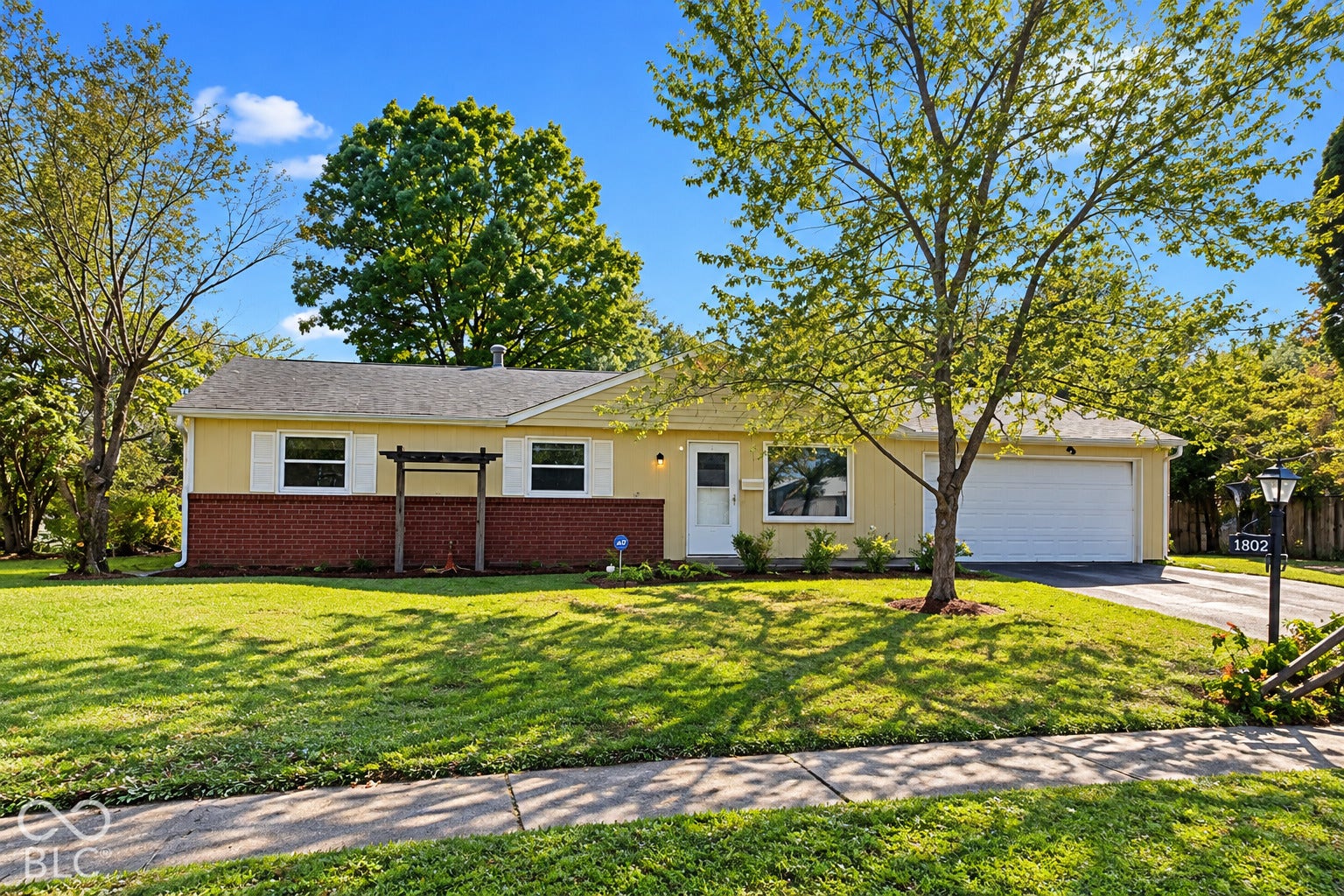 1802 Heather Court, Indianapolis, IN 46229 Virtually Altered - Ranch-style exterior featuring a mix of yellow horizontal siding and red brick accents, attached two-car garage, white window shutters, and a wooden trellis
