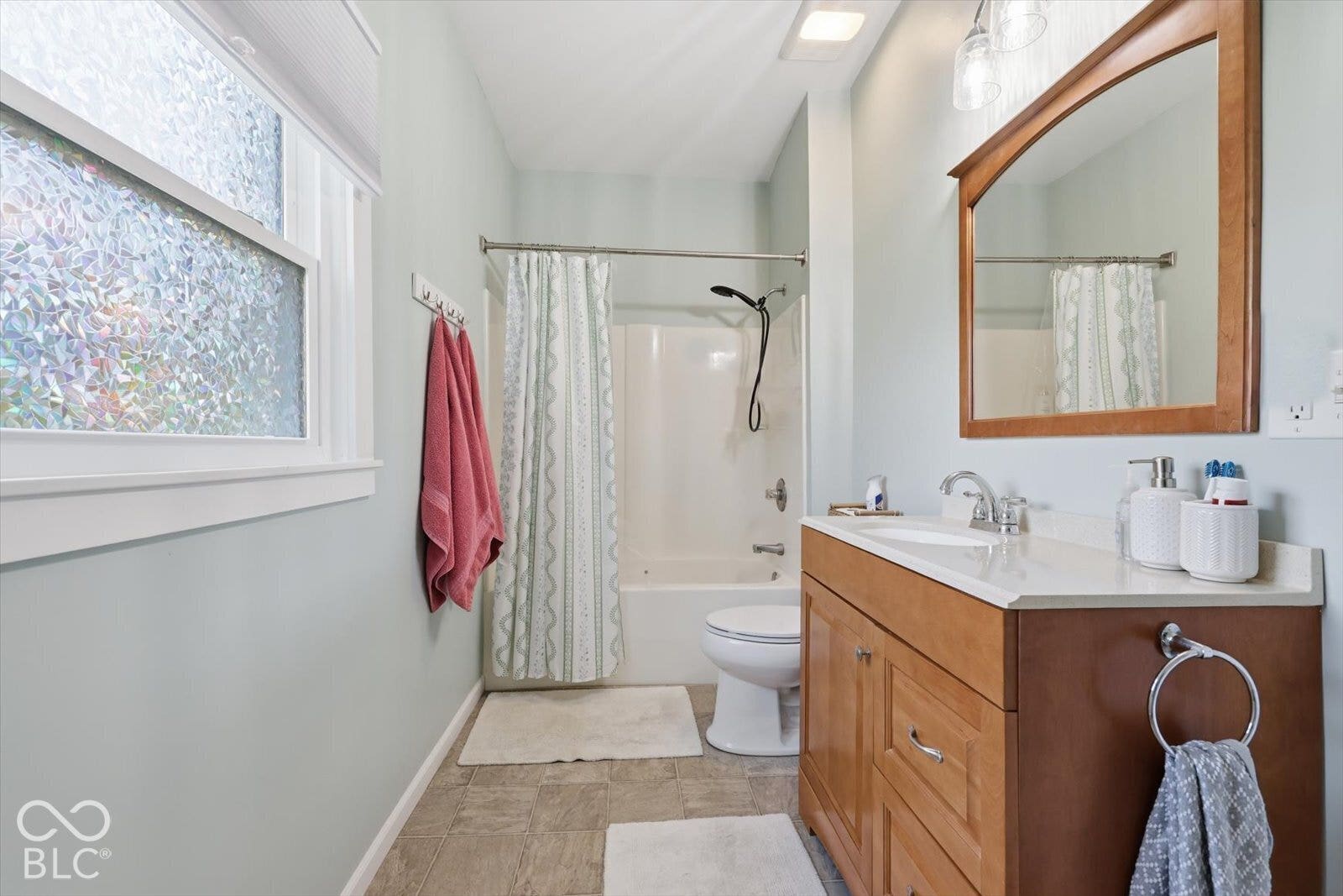 Bathroom featuring a wood-finish vanity with a stone-composite countertop, integrated sink, framed wall mirror, and a window with privacy glass
