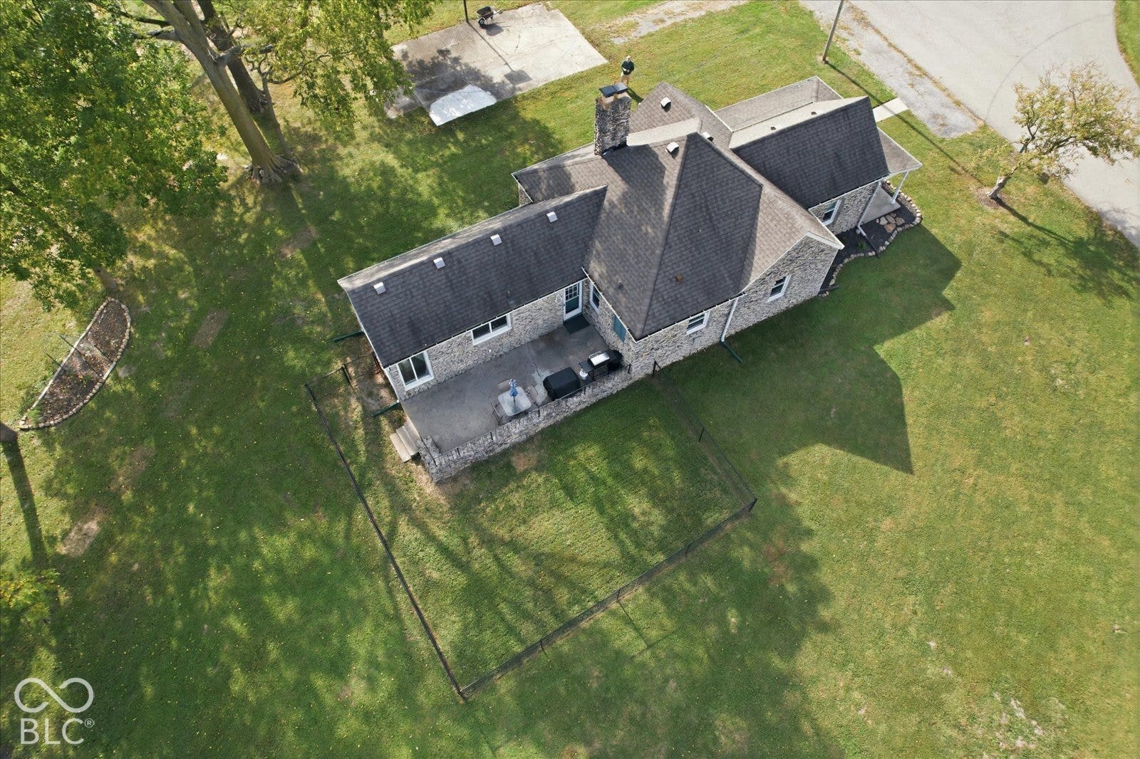 Aerial perspective of a stone-exterior residence featuring an expansive lawn, a paved concrete patio, a chimney, and a fenced-in rear yard area