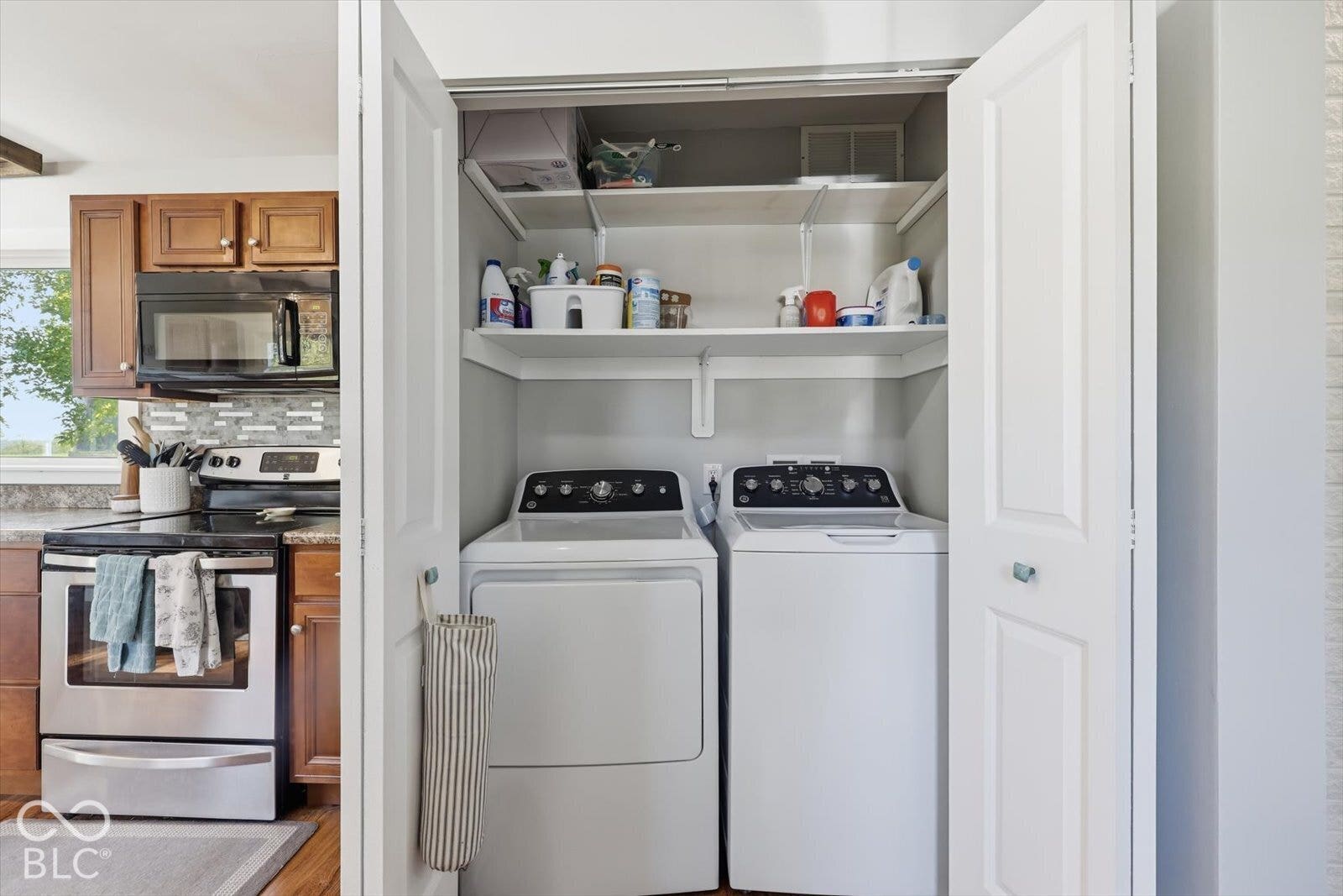 Laundry closet featuring bi-fold doors and integrated shelving, positioned near a kitchen with stainless steel appliances and wood-finish cabinetry