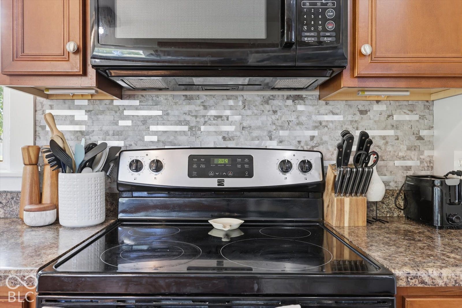 Kitchen featuring wood cabinetry, a glass-top electric range, over-the-range microwave, stone-mosaic tile backsplash, and solid-surface countertops