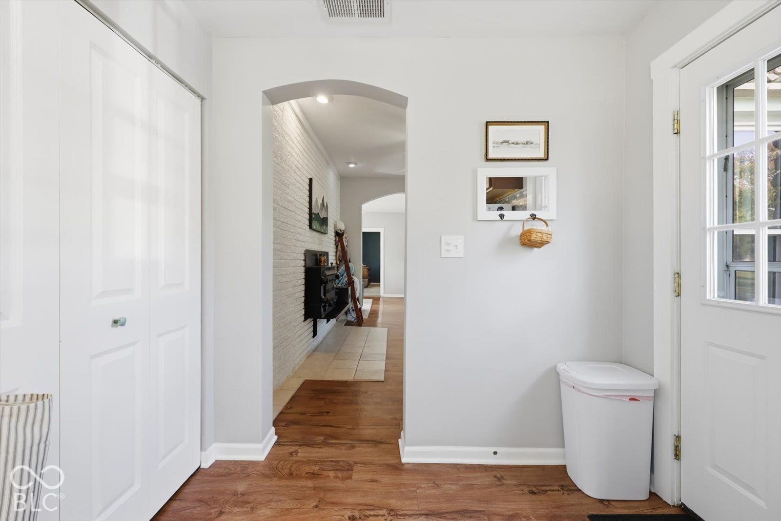 Entryway featuring wood-finish flooring, a multi-panel door with glass inserts, white bifold closet doors, and a decorative arched wall opening revealing a white painted brick wall