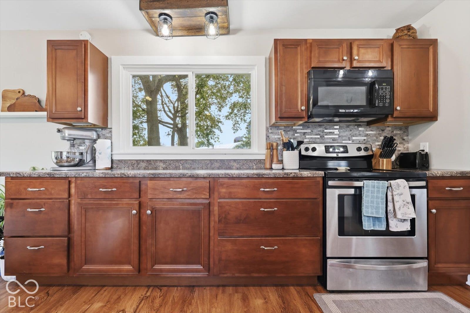 Kitchen featuring stained wood cabinetry, stone-finish countertops, a multi-tonal linear tile backsplash, stainless steel appliances, and a rectangular ceiling-mounted light fixture with glass shades