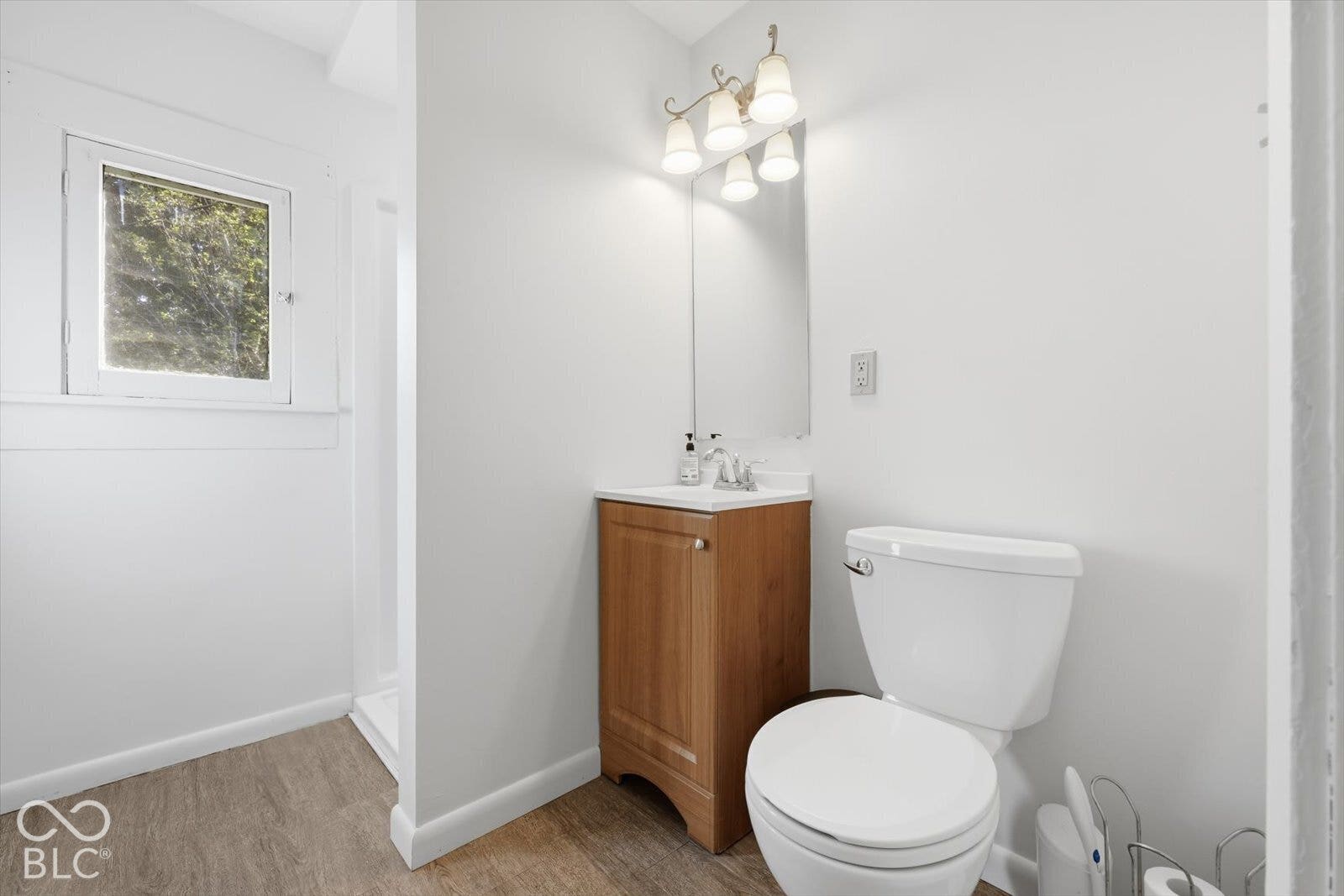 Bathroom featuring wood-finish flooring, a vanity with a white countertop and sink, a wall-mounted mirror, four-light vanity fixture, and a white toilet
