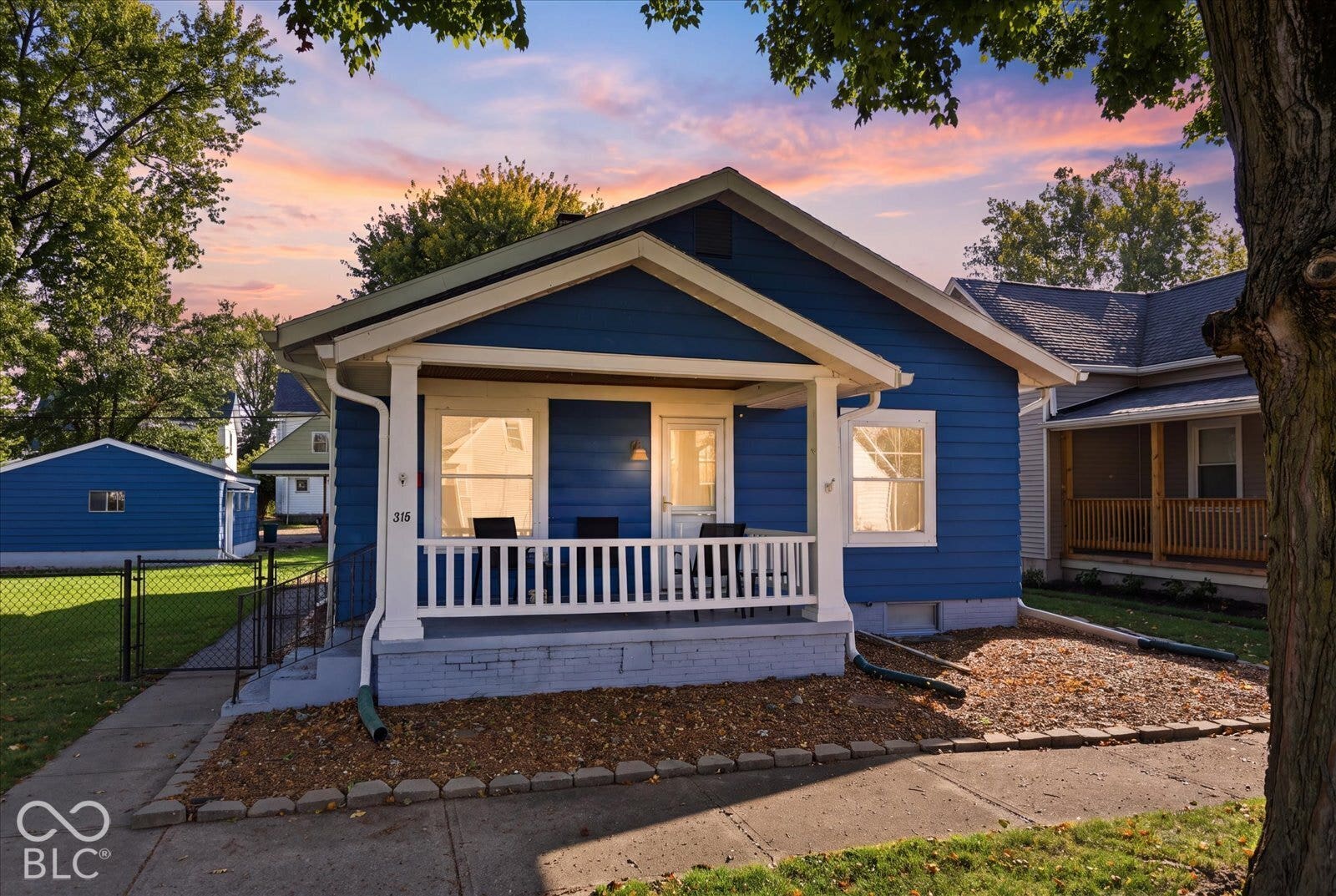 Virtually Altered - Blue siding exterior featuring a covered front porch with white railing, brick foundation, and a detached structure with a fenced yard