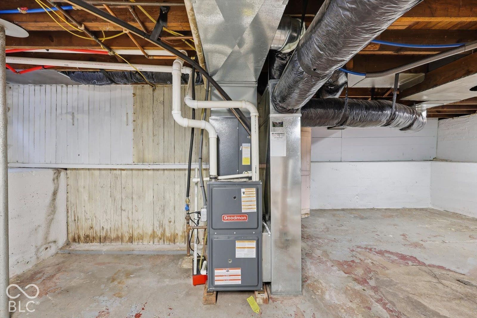 Goodman forced-air furnace unit featuring a metal ductwork system, PVC plumbing lines, concrete flooring, and painted masonry walls