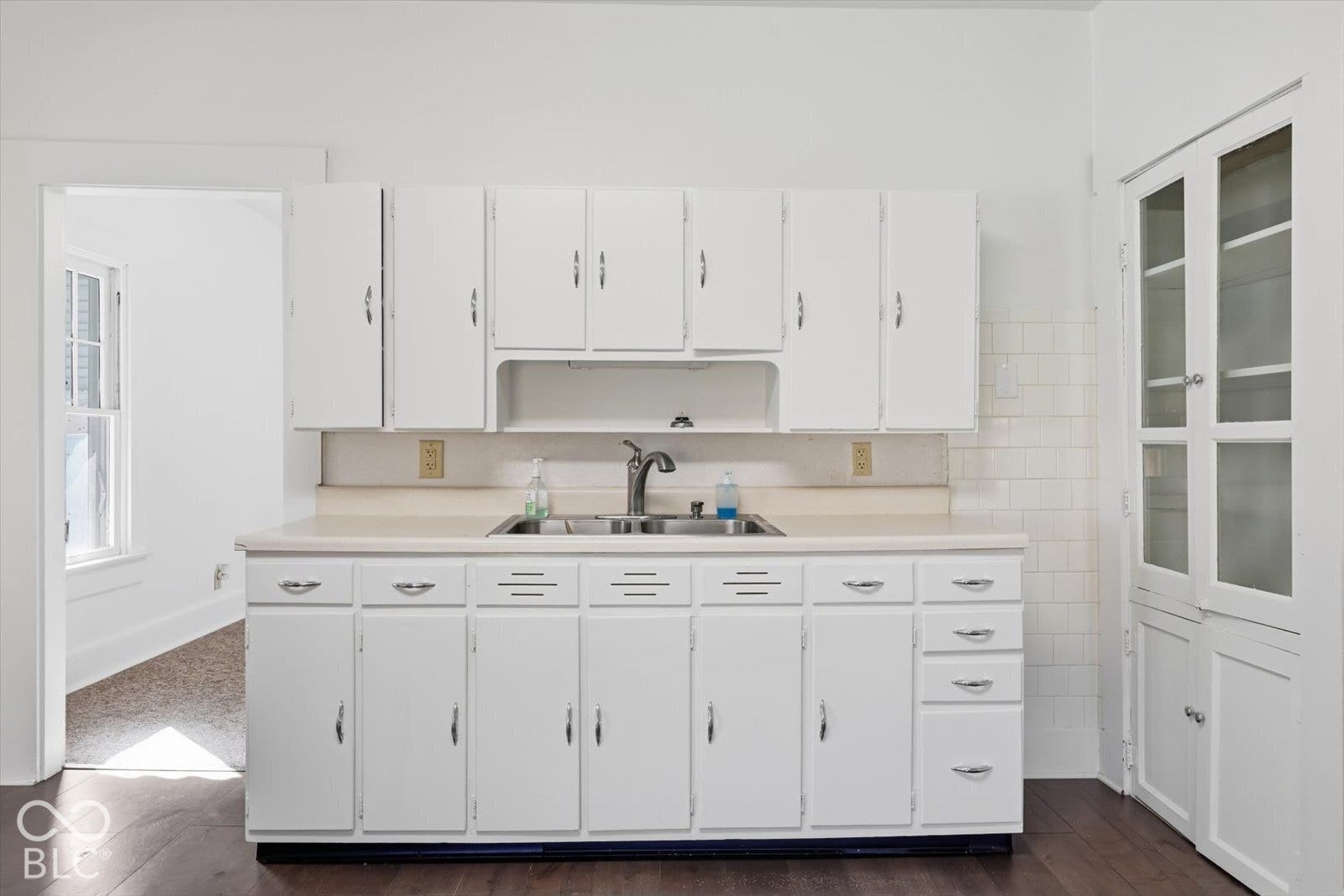Kitchen featuring white shaker-style cabinetry, integrated glass-front display hutch, double-basin stainless steel sink, gooseneck faucet, and tile backsplash