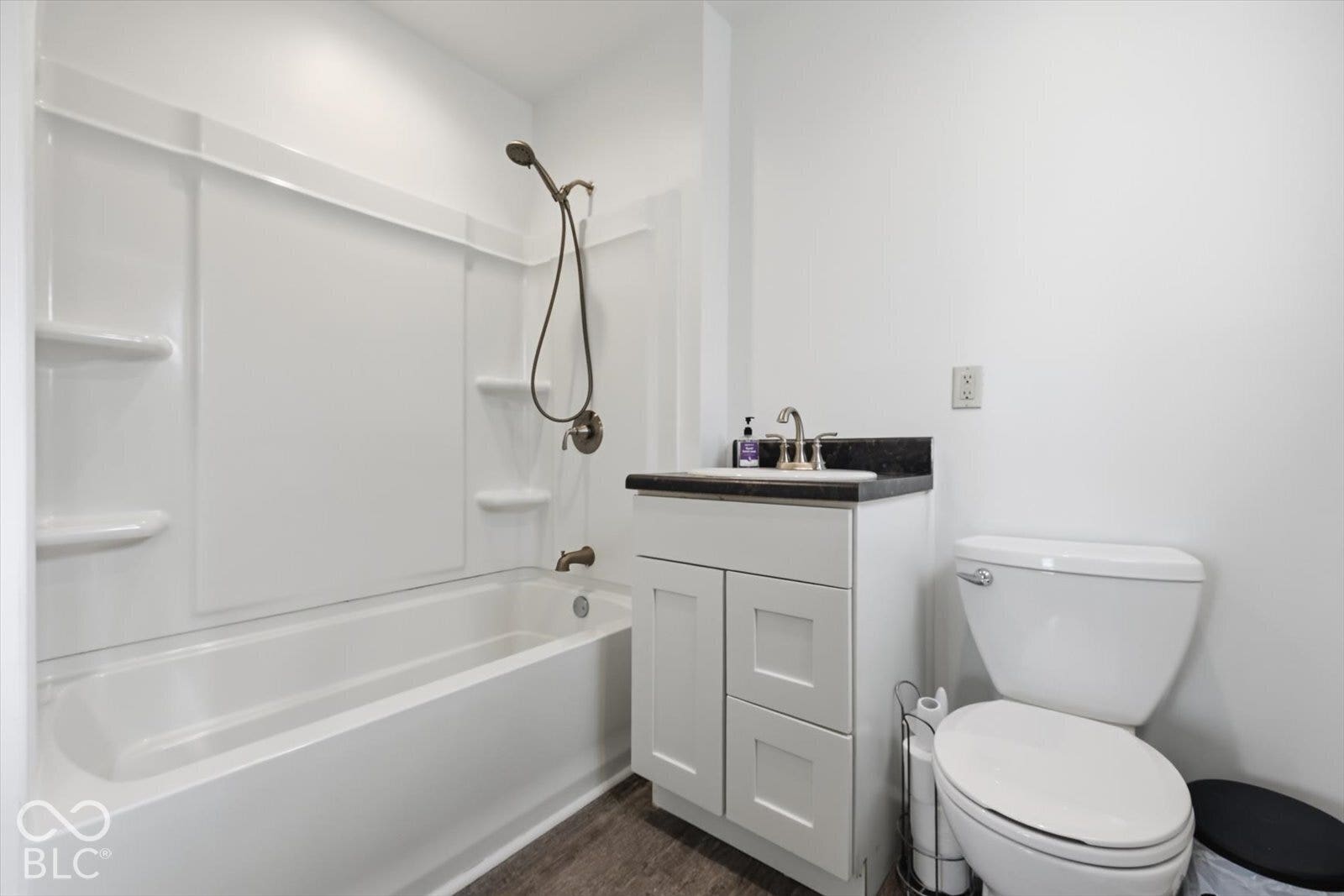 Bathroom featuring a shower-tub combination with integrated shelving, a vanity with a dark stone-finish countertop and undermount sink, and wood-finish flooring