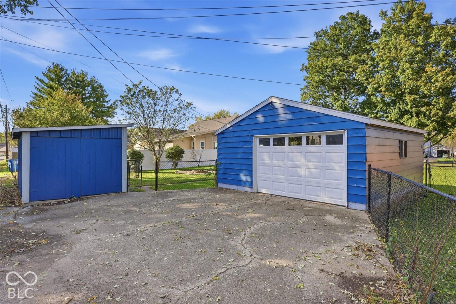Detached two-car garage featuring a white sectional door, auxiliary storage structure, and concrete parking area enclosed by chain-link fencing