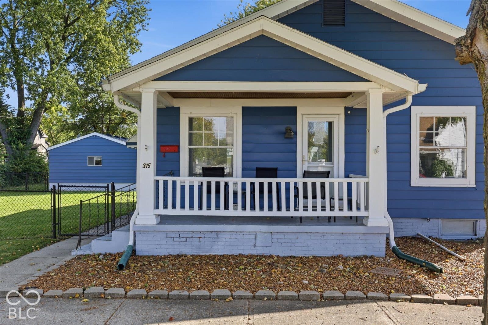 Blue exterior siding featuring a covered front porch, white railings, a detached garage, and a chain-link fenced yard