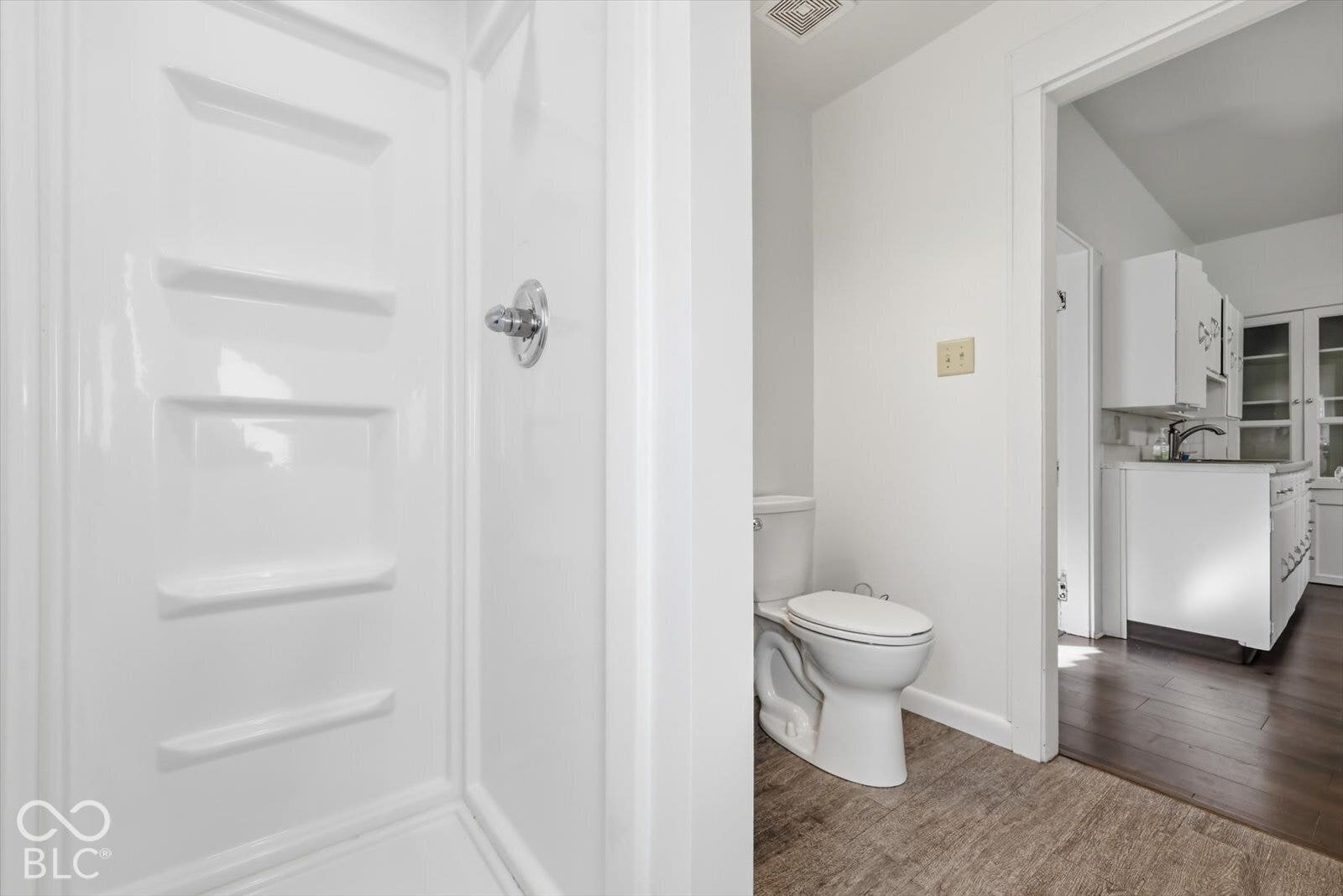 Bathroom featuring a white shower surround with integrated shelving, chrome shower hardware, and a standard-height toilet, with wood-finish flooring extending into the room