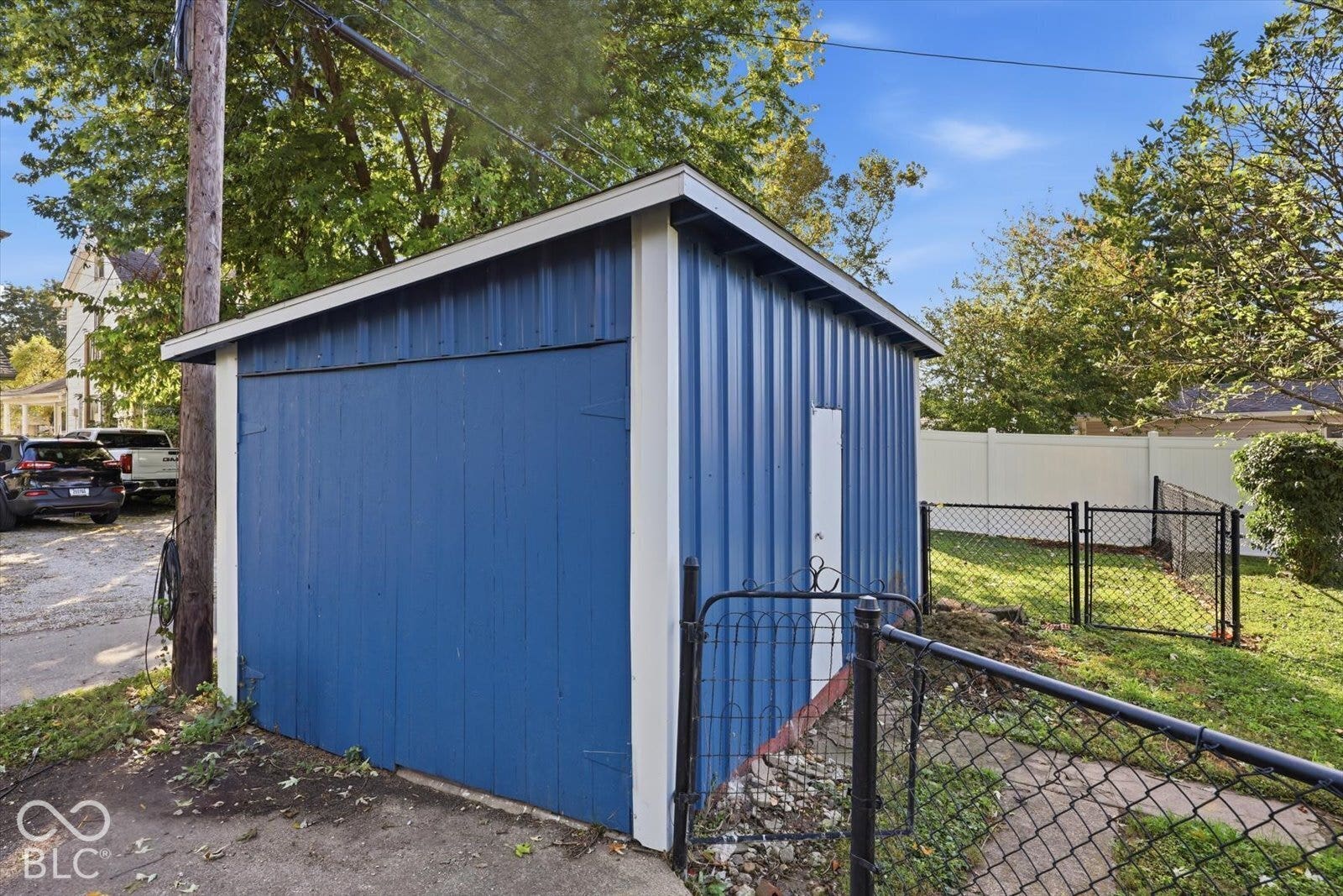 Detached storage structure featuring blue vertical metal siding, white trim, a single-door entrance, and a chain-link fenced perimeter