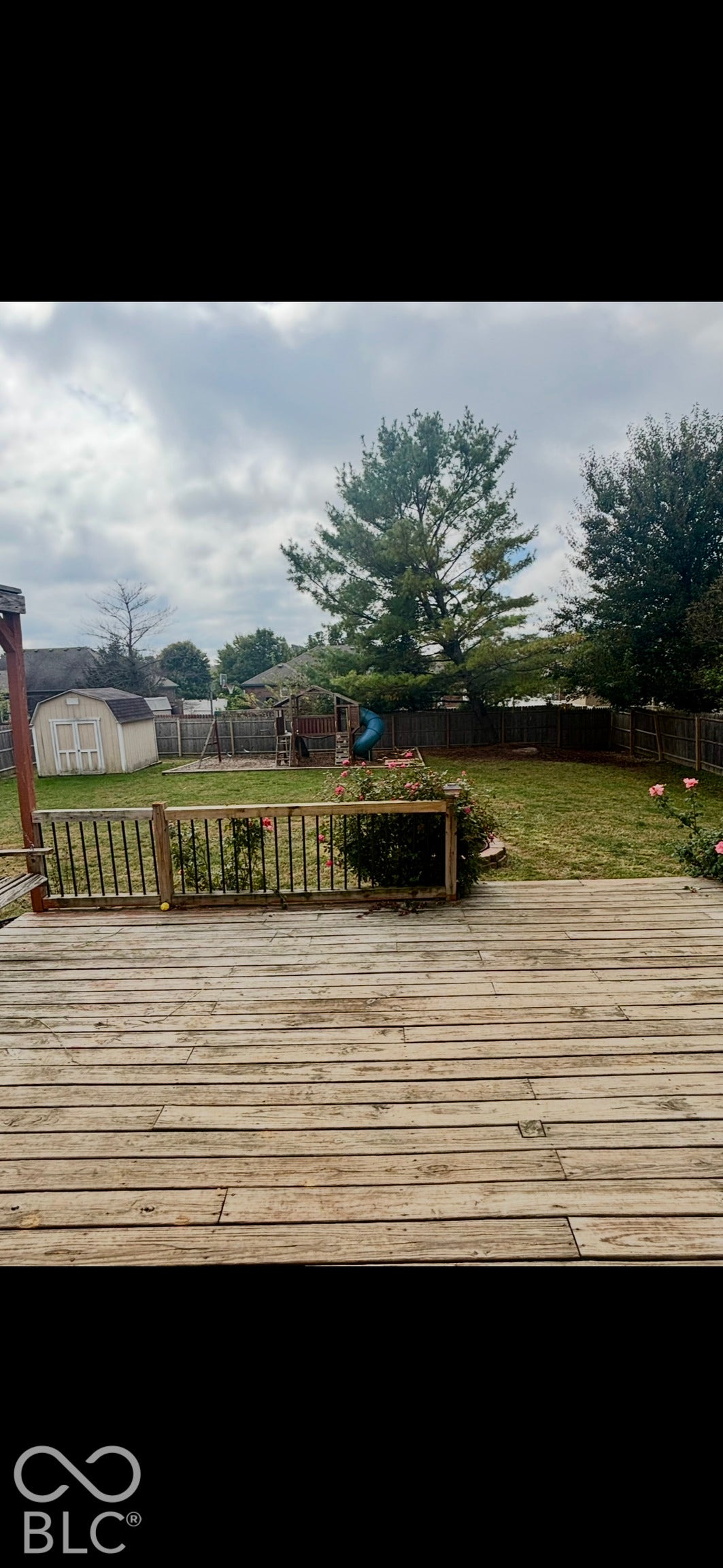 Rear wood deck featuring a baluster railing, expansive fenced-in lawn, mature trees, and an accessory storage structure