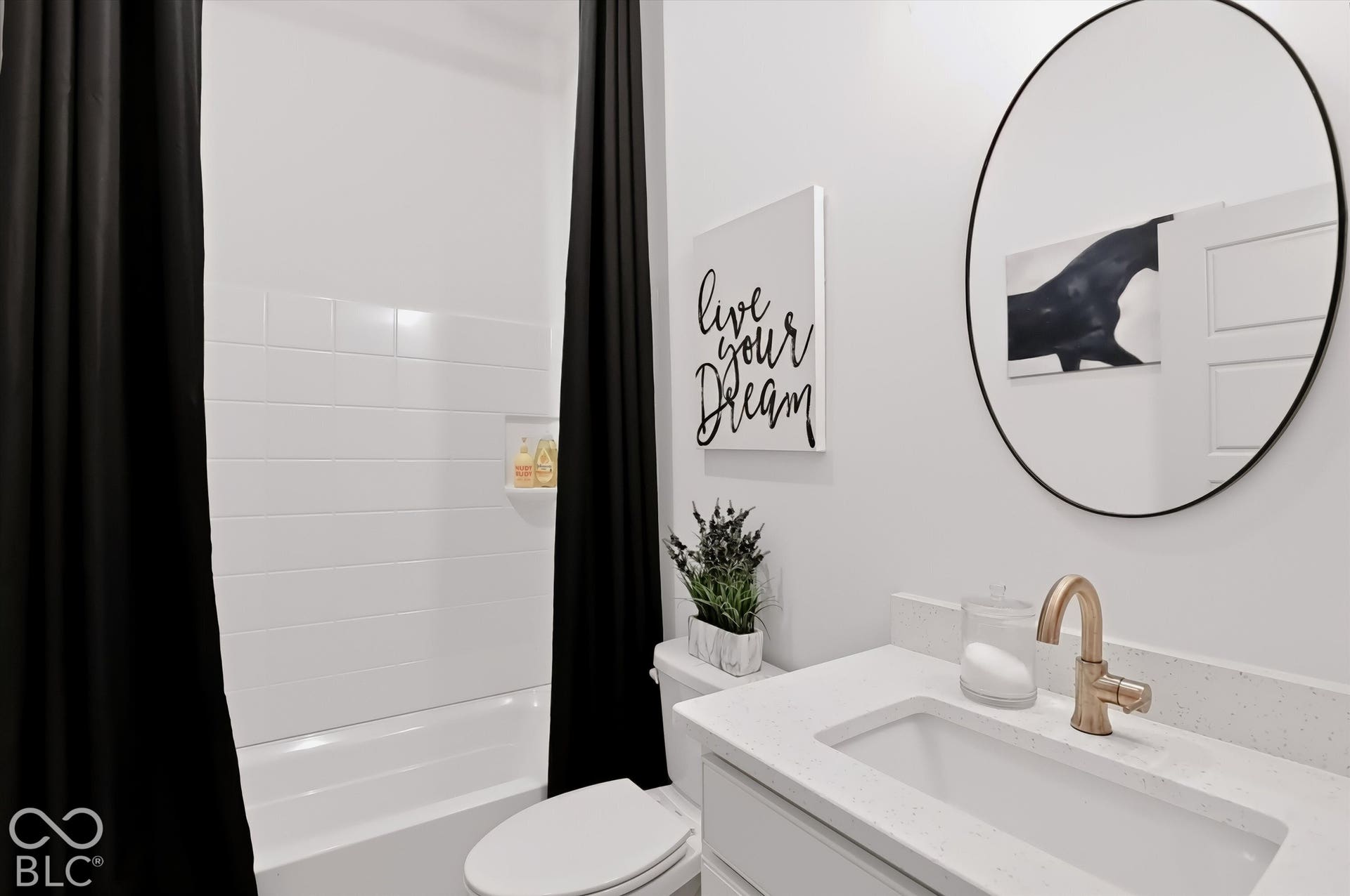 Bathroom featuring a vanity with a white composite countertop, an undermount sink, and a metallic finish faucet, complemented by an oval wall-mounted mirror, white wall tile surround, and an integrated shower niche