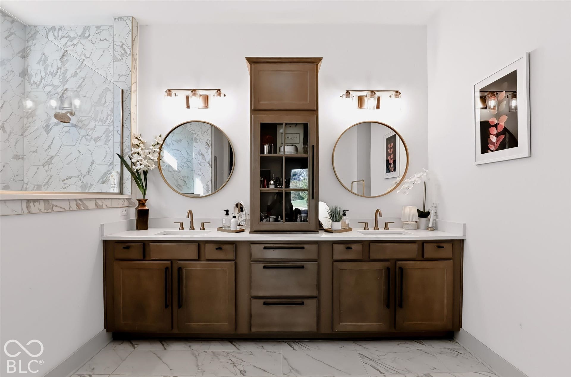Dual-sink vanity featuring stained wood cabinetry with built-in center tower, stone-surface countertop, two circular wall-mounted mirrors, and tiled shower enclosure