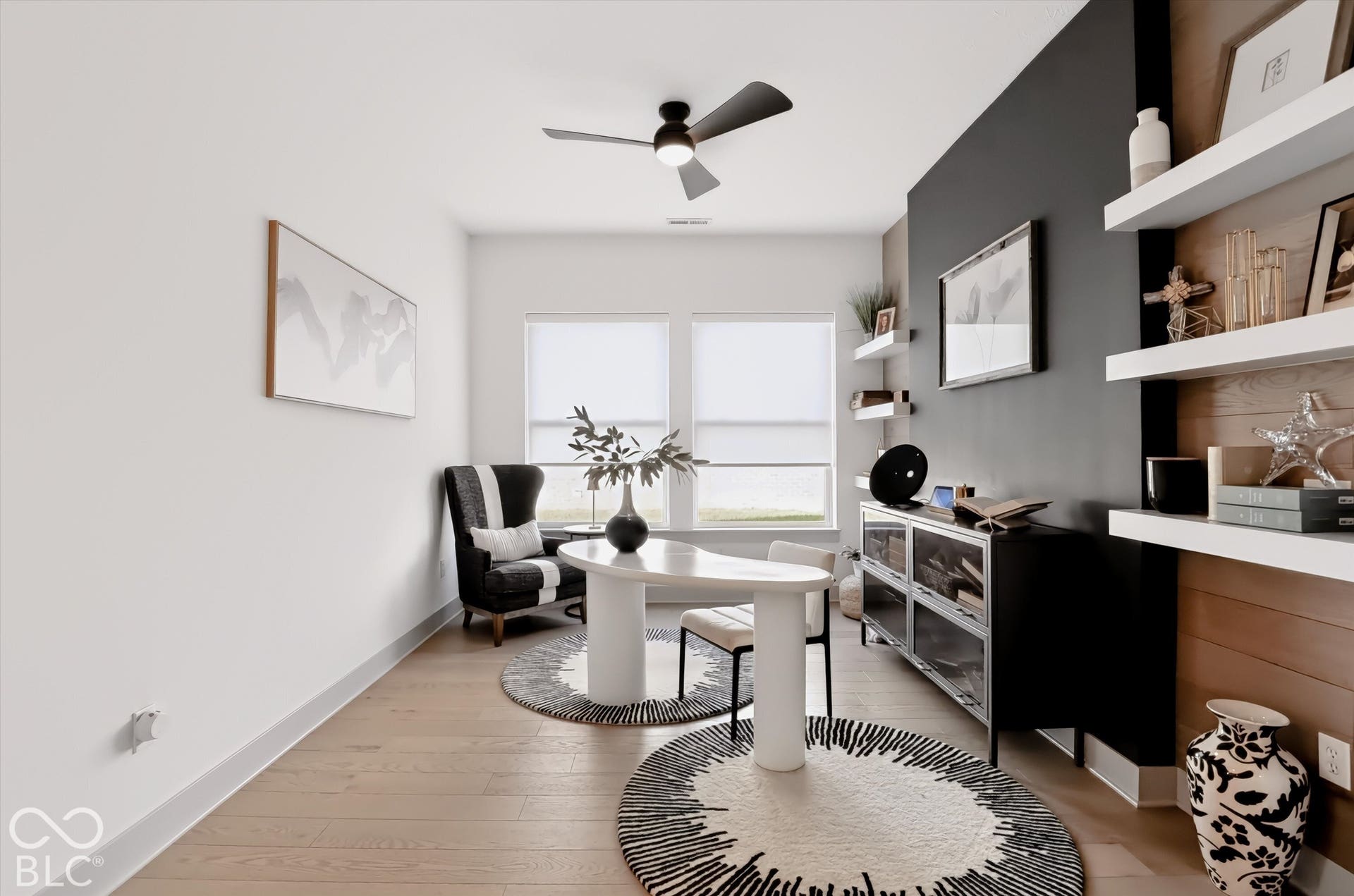 Wood-finish flooring, dual windows with roller shades, a ceiling fan with integrated lighting, a charcoal-toned accent wall, and wall-mounted floating shelves