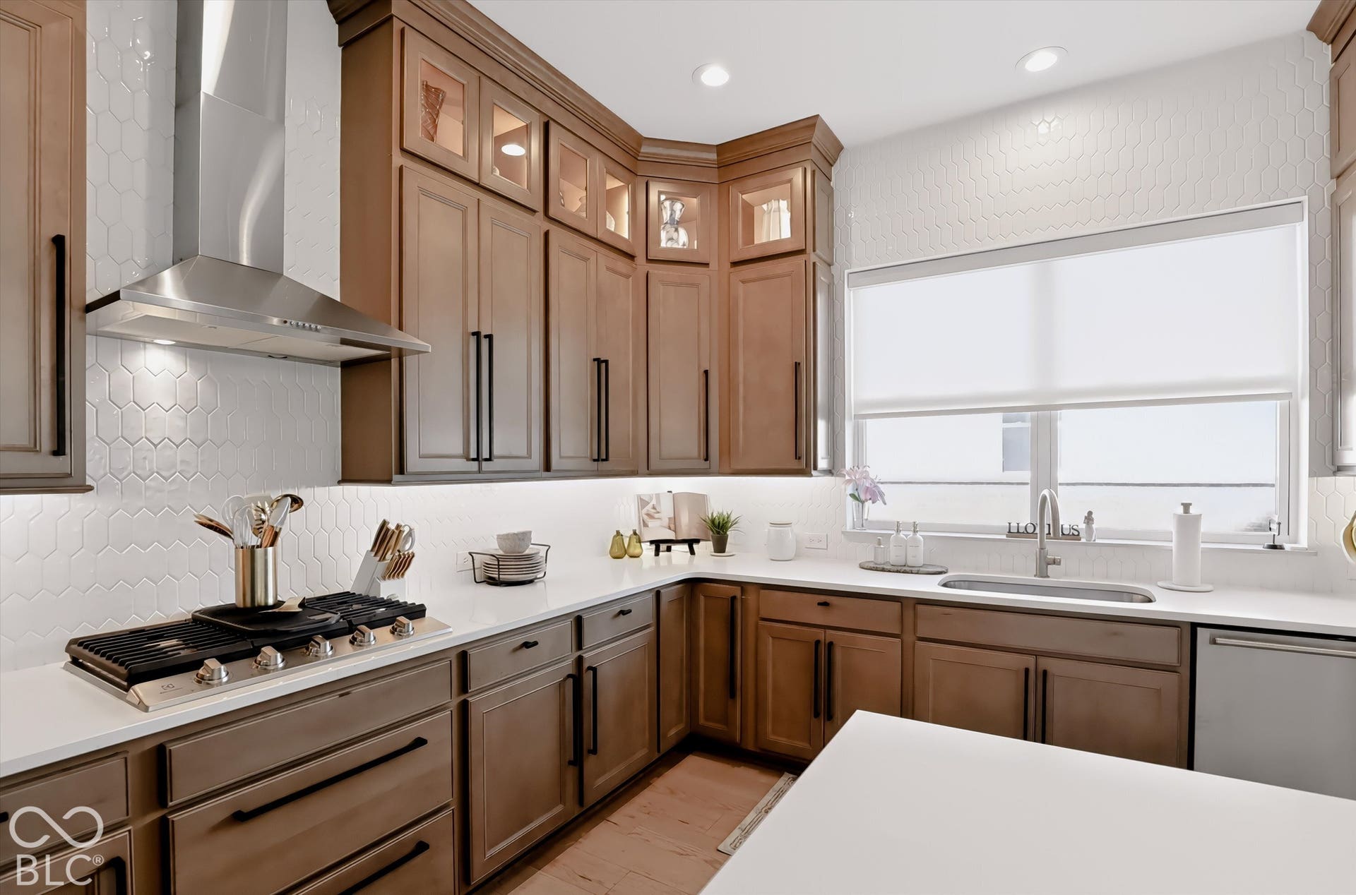 Kitchen featuring wood cabinetry with glass-front uppers, solid-surface countertops, a white hexagonal tile backsplash, a stainless steel gas cooktop with matching range hood, and wood-finish flooring
