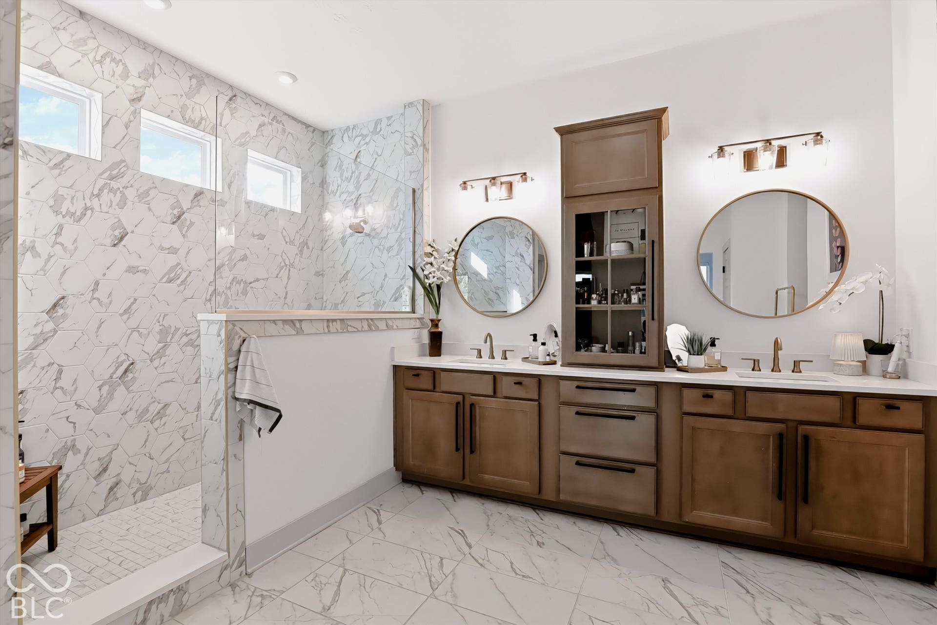 Dual-vanity cabinetry with a central built-in storage tower, stone-finish tile flooring, walk-in shower with floor-to-ceiling stone-finish surround, and circular wall-mounted mirrors