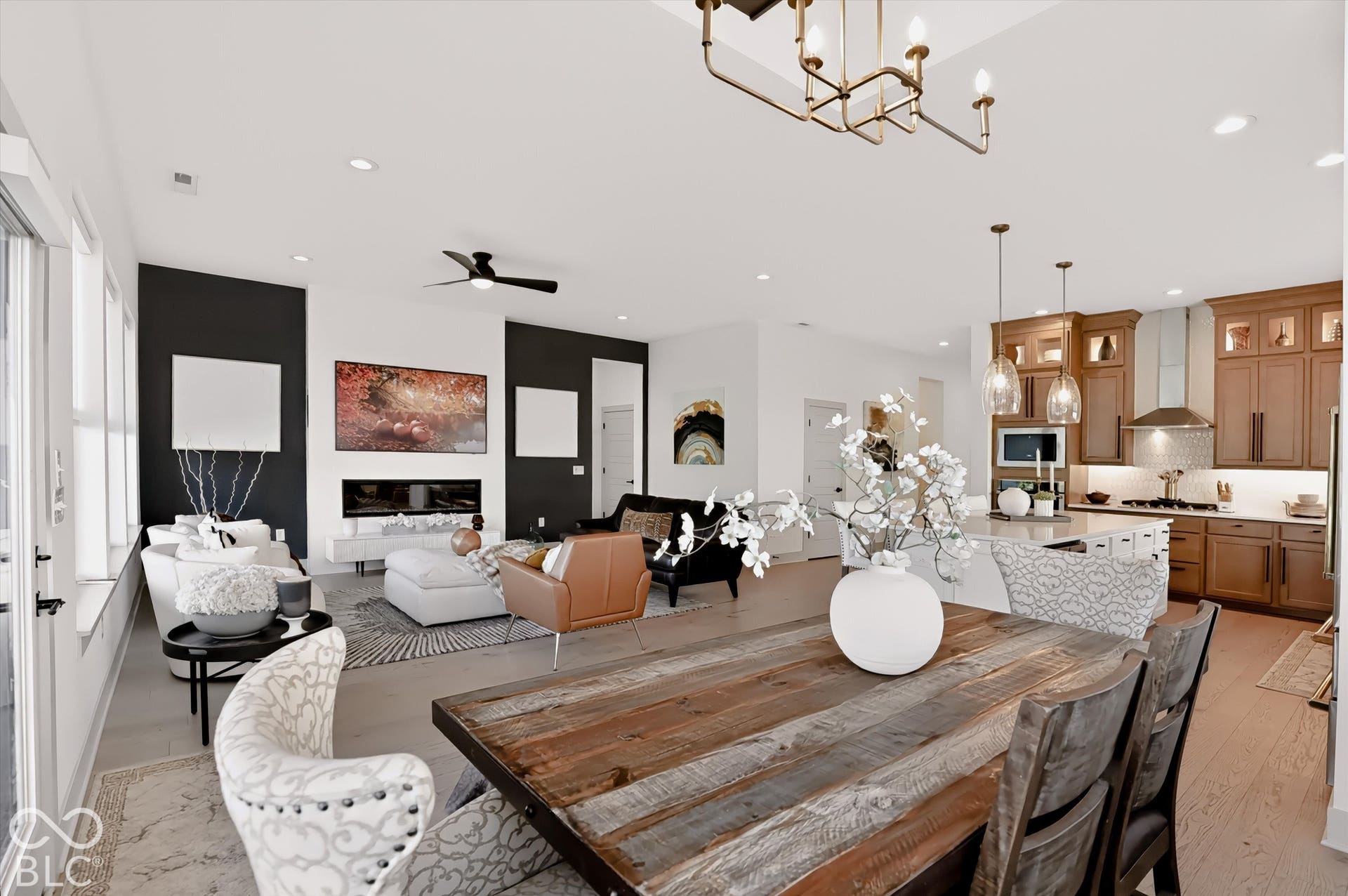 Open-concept living space featuring wood-finish flooring, a contemporary linear fireplace with a high-contrast accent wall, natural wood cabinetry, a center kitchen island with pendant lighting, and recessed ceiling lights