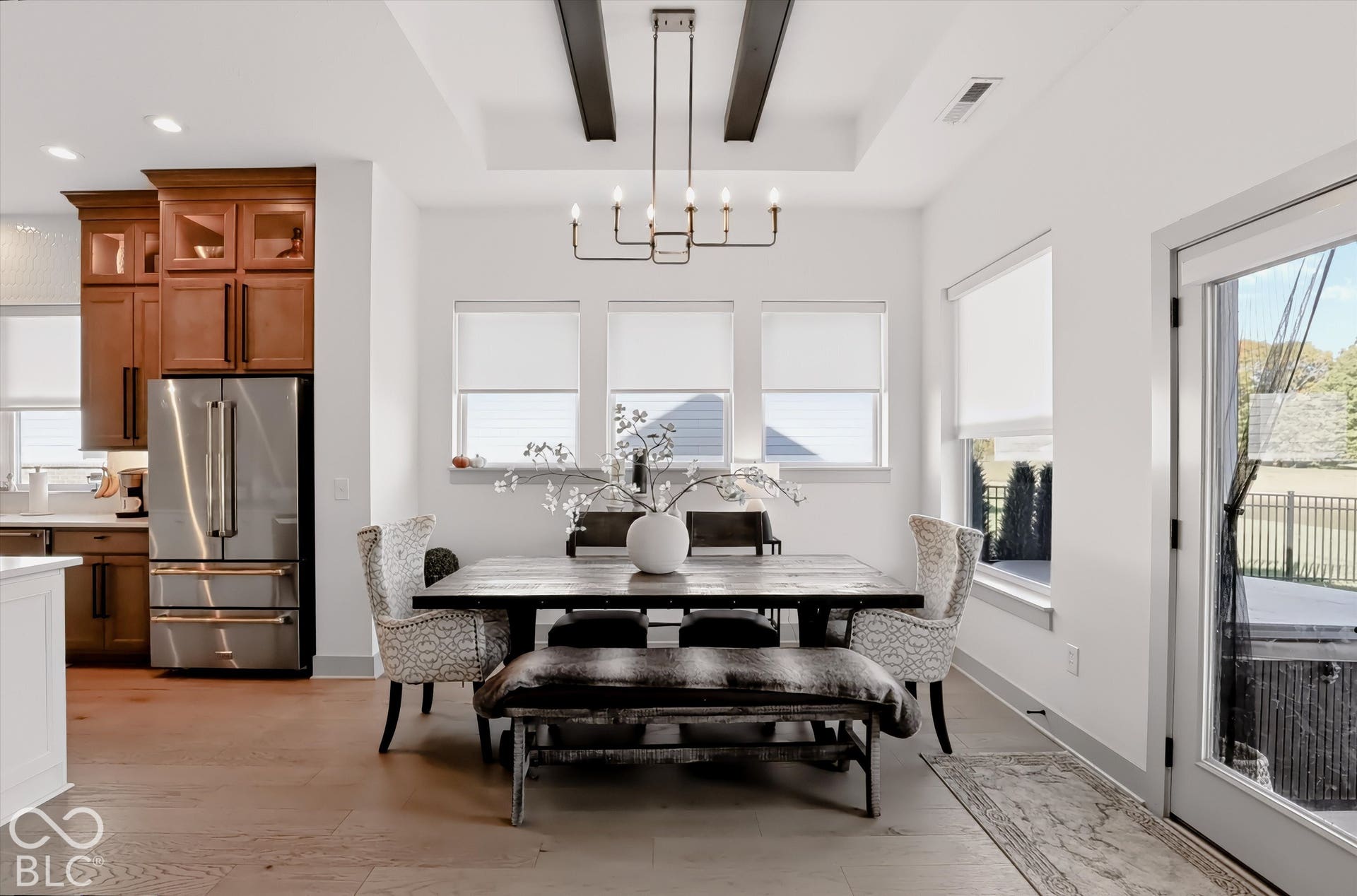 Tray ceiling with dark wood beams, wood-finish flooring, custom wood-tone cabinetry, stainless steel refrigerator, and industrial-style multi-light chandelier