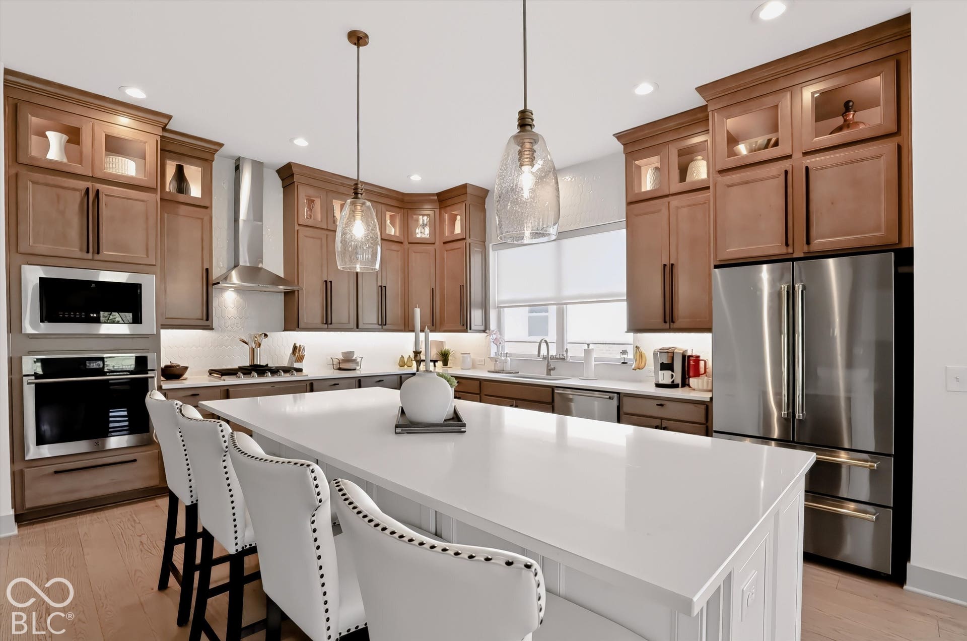Kitchen featuring stained wood cabinetry with glass-front uppers, a central island with a solid-surface countertop, stainless steel appliances including a built-in oven and microwave, recessed lighting, and pendant fixtures
