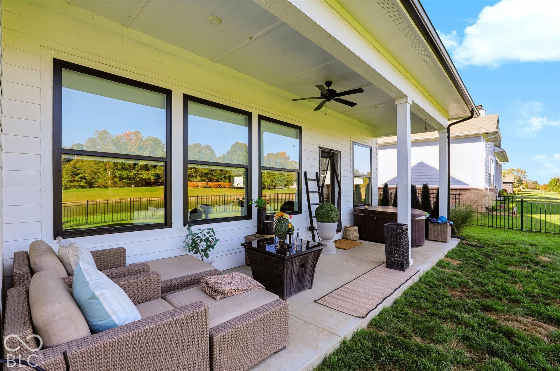 Covered concrete patio featuring white horizontal siding, large windows with black frames, a ceiling-mounted fan, and a perimeter metal fence