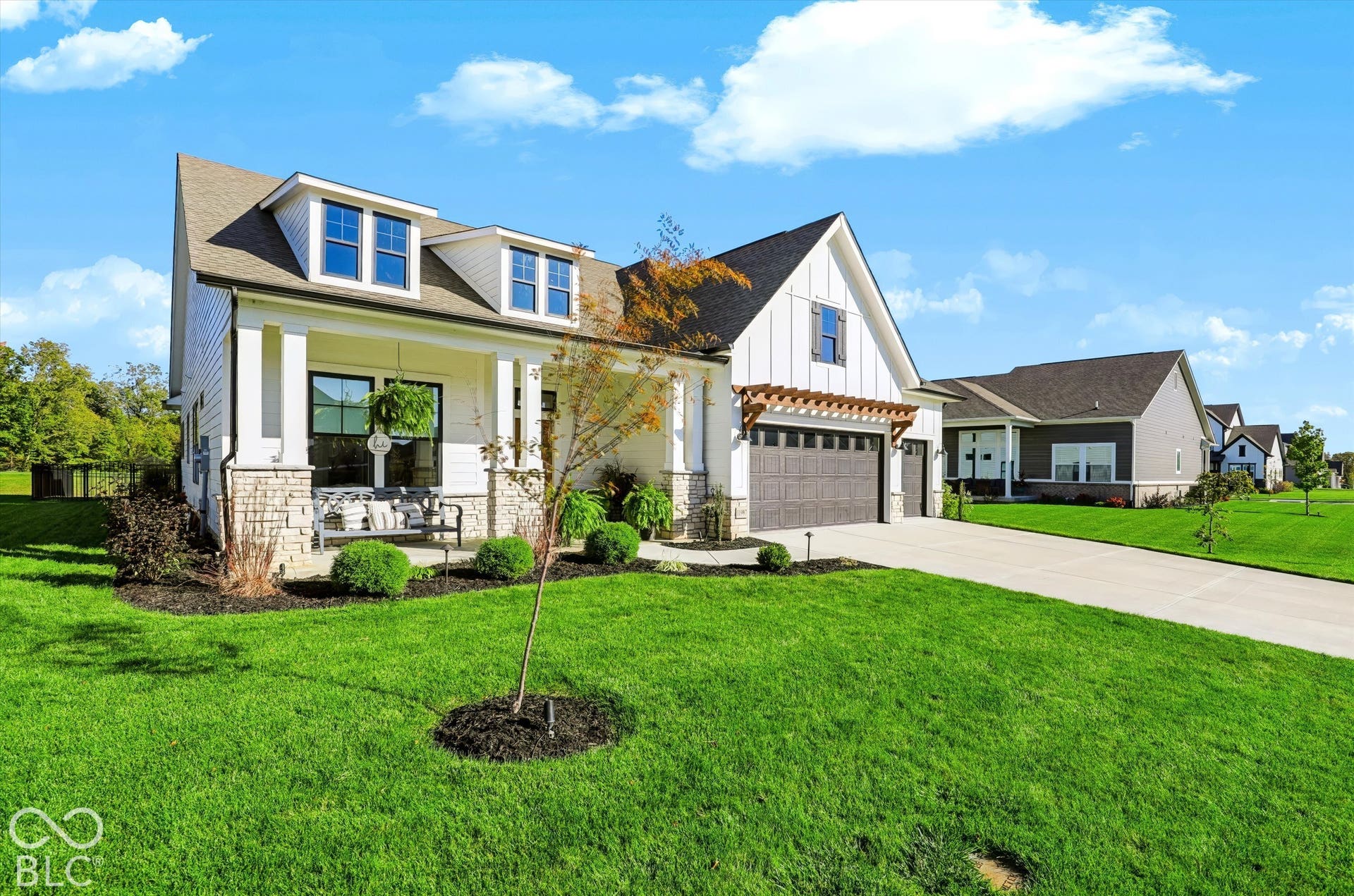 Modern farmhouse exterior featuring white board-and-batten siding, natural stone veneer accents, a covered front porch with square columns, a dark-toned multi-car garage with a decorative wood pergola, and dormer windows
