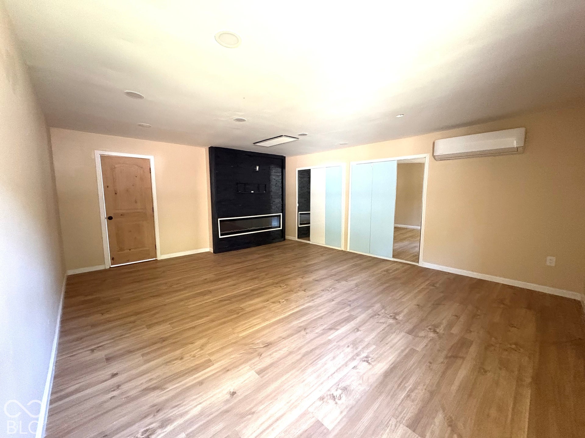 Wood-finish flooring, black tiled accent wall with integrated linear fireplace, dual sliding-mirror closets, and wall-mounted ductless climate control unit