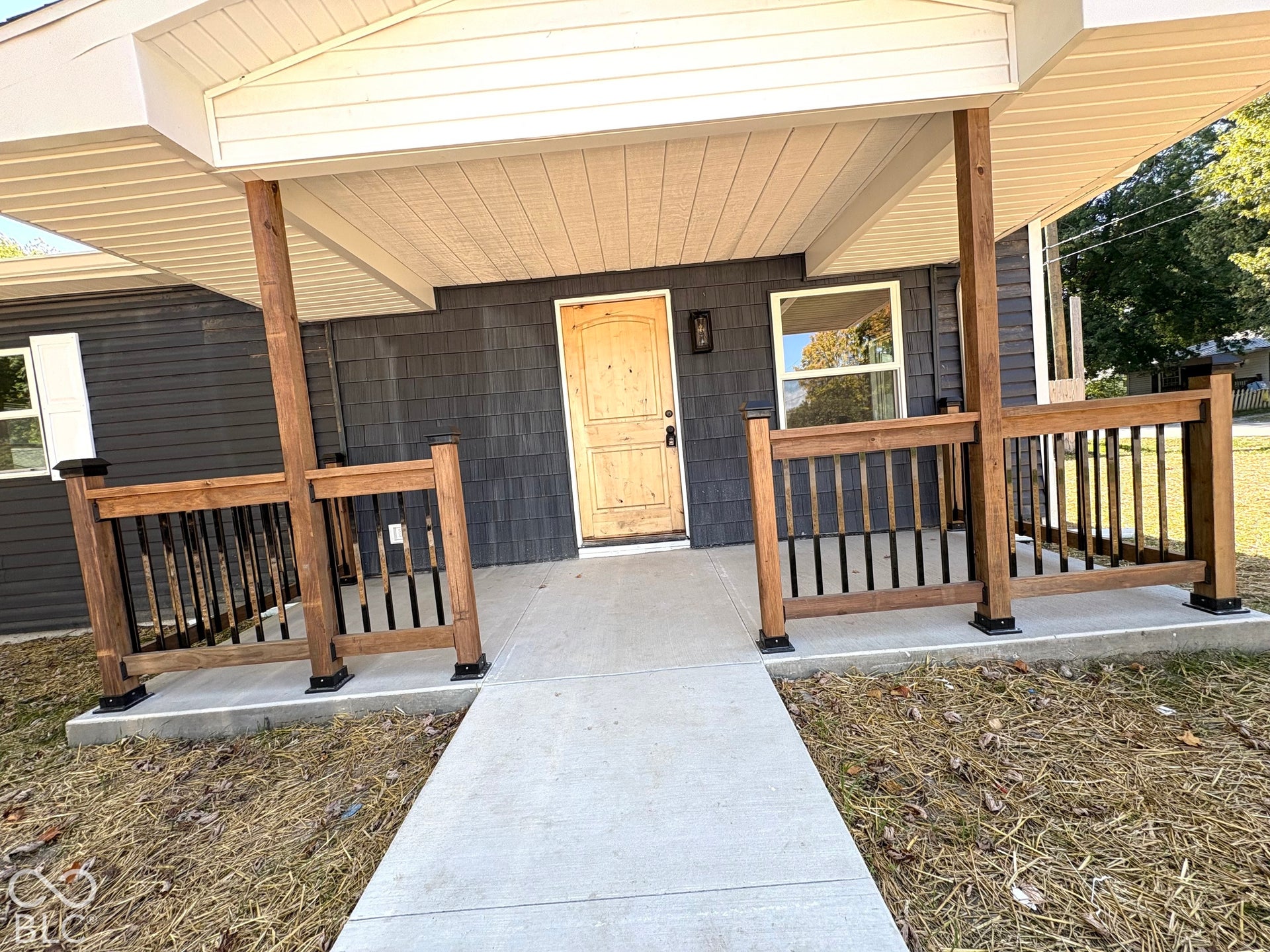 Covered front porch featuring dark-tone shake siding, a natural wood-finish front door, and a concrete entry path with wood-post railings and black balusters