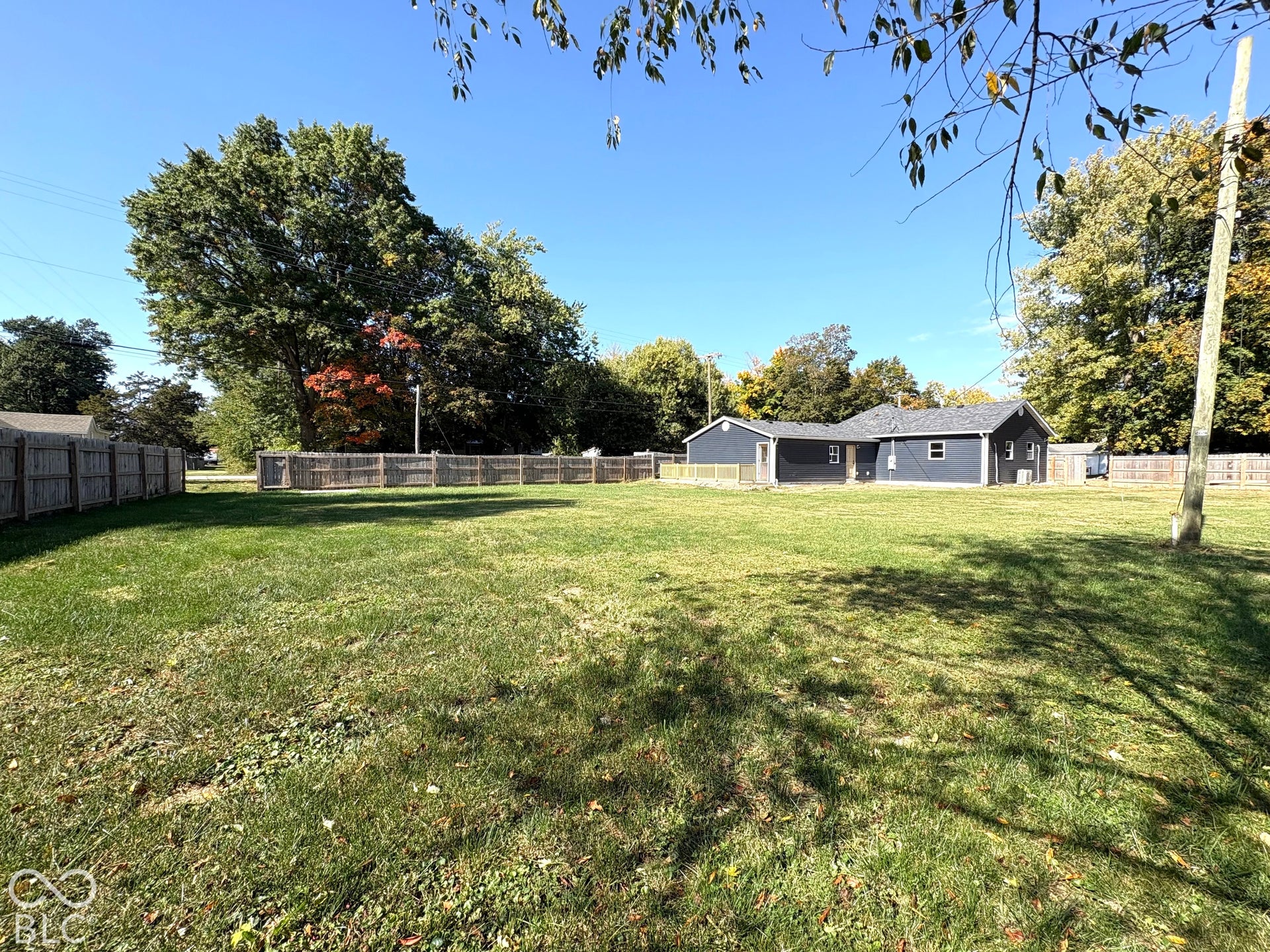 Expansive level backyard featuring a perimeter wood privacy fence, mature trees, and a dark-toned exterior structure with an attached deck