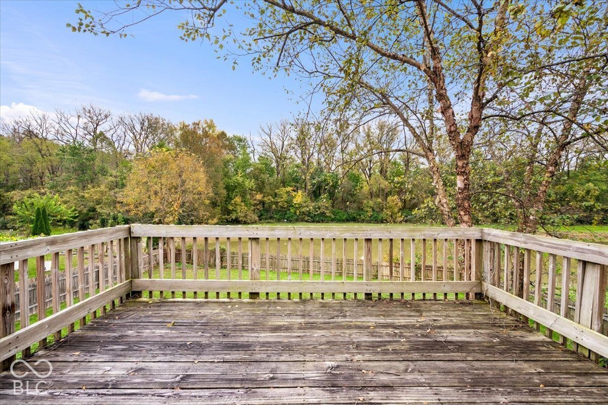 Elevated wood deck featuring a balustrade railing, overlooking a fenced backyard with mature trees