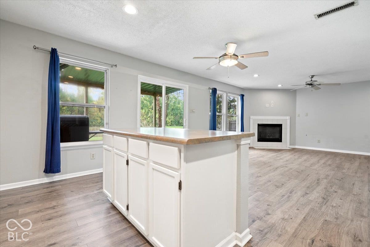 Open-concept interior featuring wood-finish flooring, a kitchen island with stone-finish countertop, dual ceiling fans, sliding glass doors, and a fireplace with a white mantle