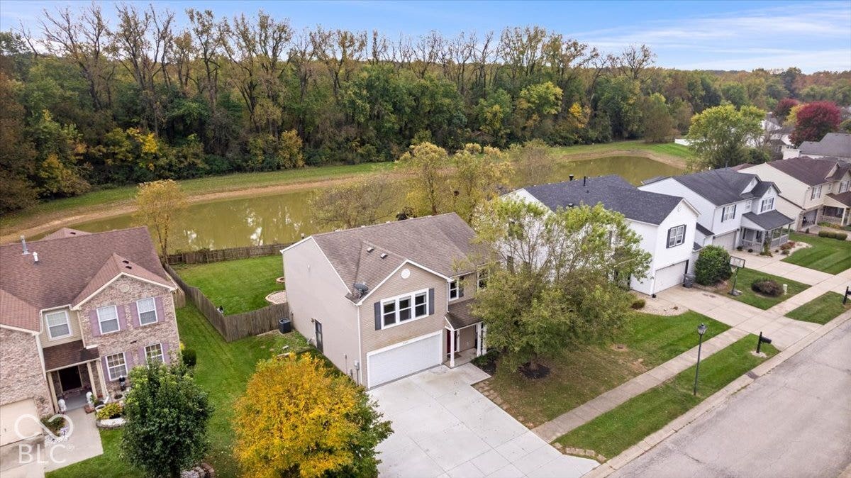 Aerial perspective featuring a two-story residence with a two-car garage, shingled roof, concrete driveway, and a fenced rear yard bordering a pond