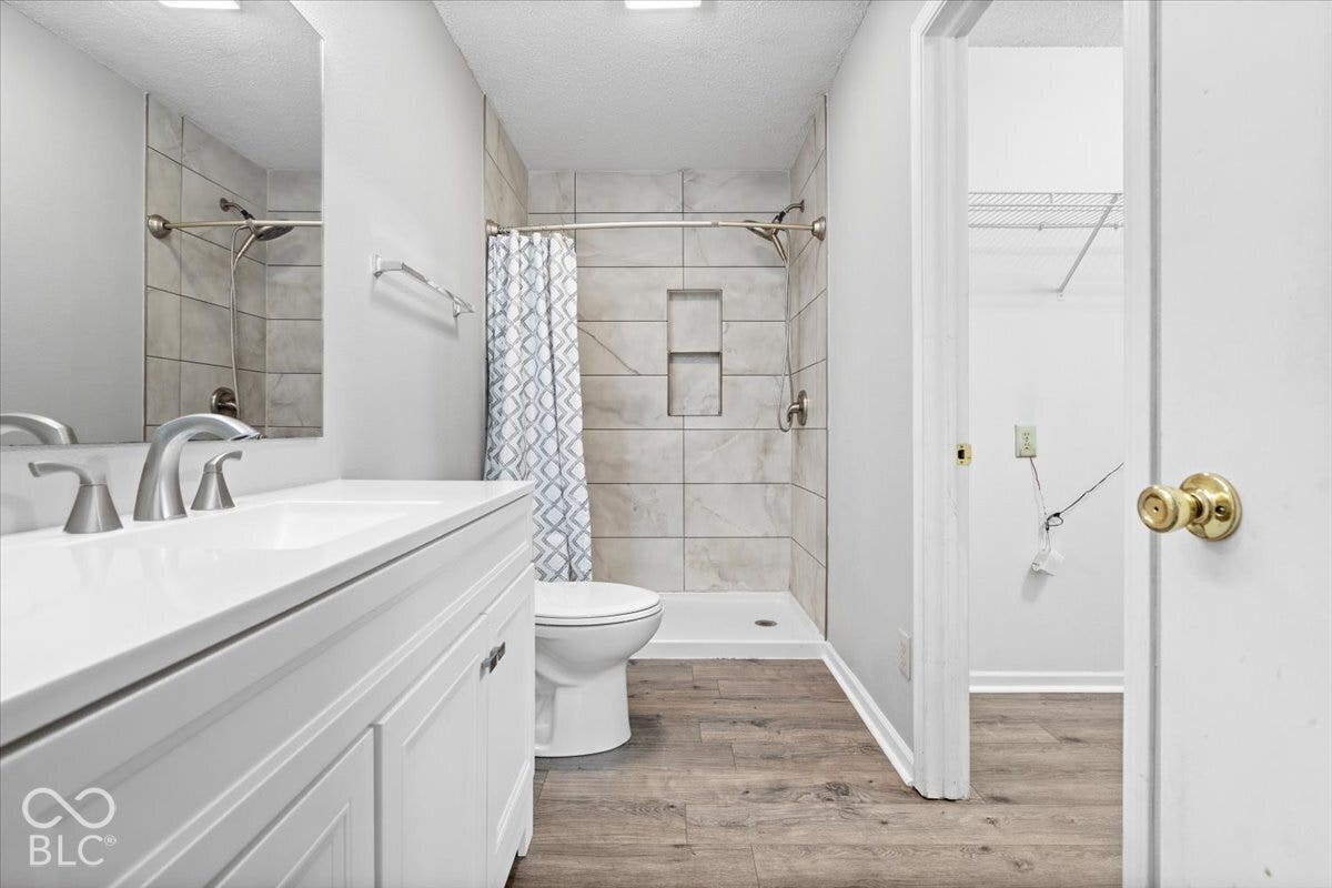 Bathroom featuring wood-finish flooring, a white vanity with a rectangular sink, large frameless mirror, and a walk-in shower with stone-style wall tile and a built-in recessed niche