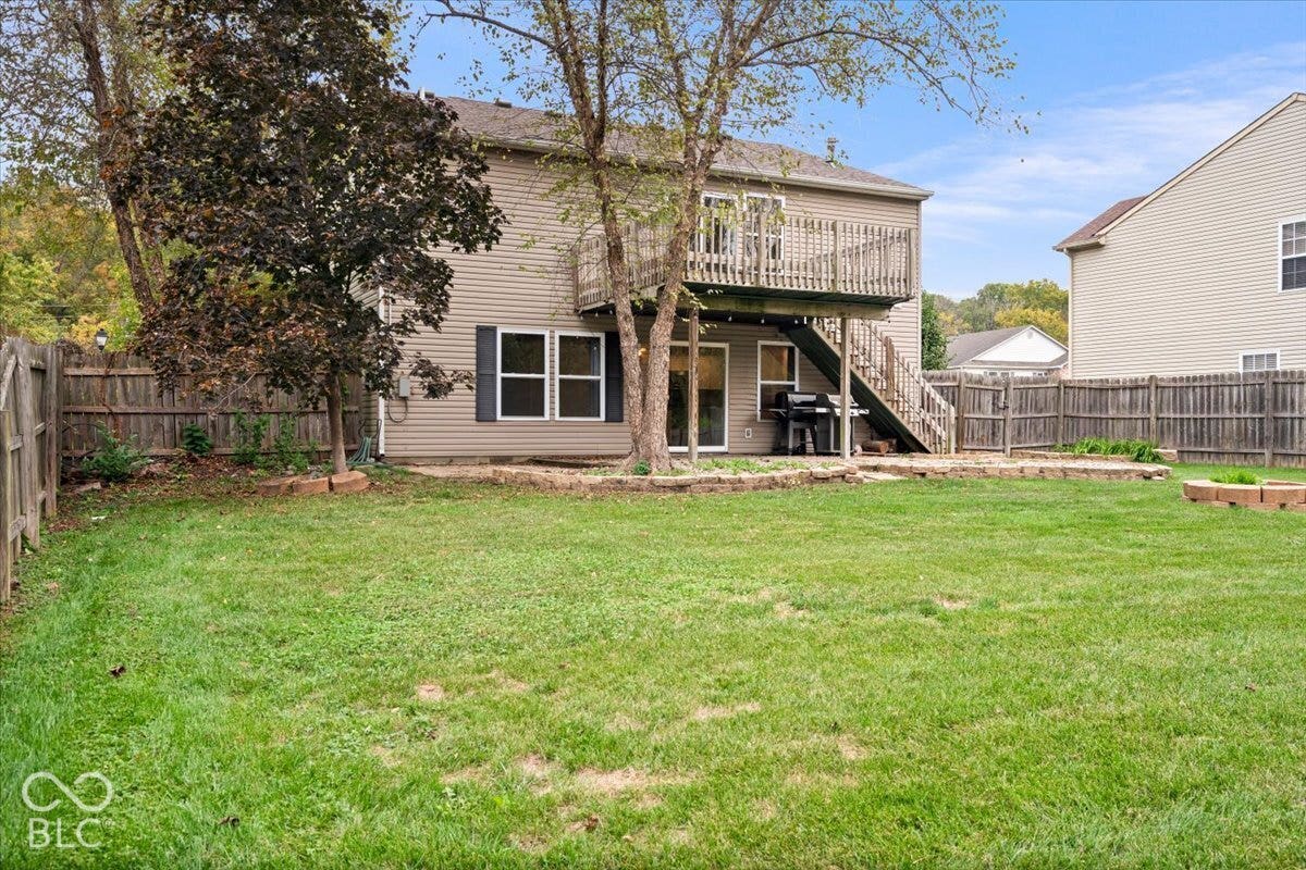 Rear exterior featuring light-toned horizontal siding, a wooden elevated deck with stairs, a stone-bordered patio, and full wood perimeter fencing