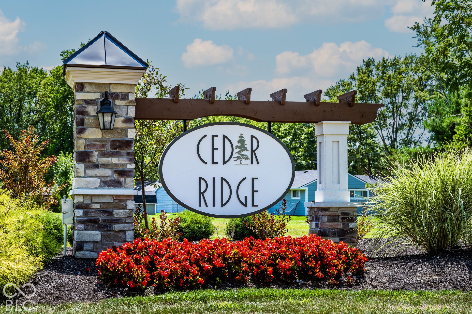 Community signage featuring stone-clad masonry columns, a wood-beam pergola, metal cap accents, and integrated lantern-style light fixtures surrounded by manicured landscaping
