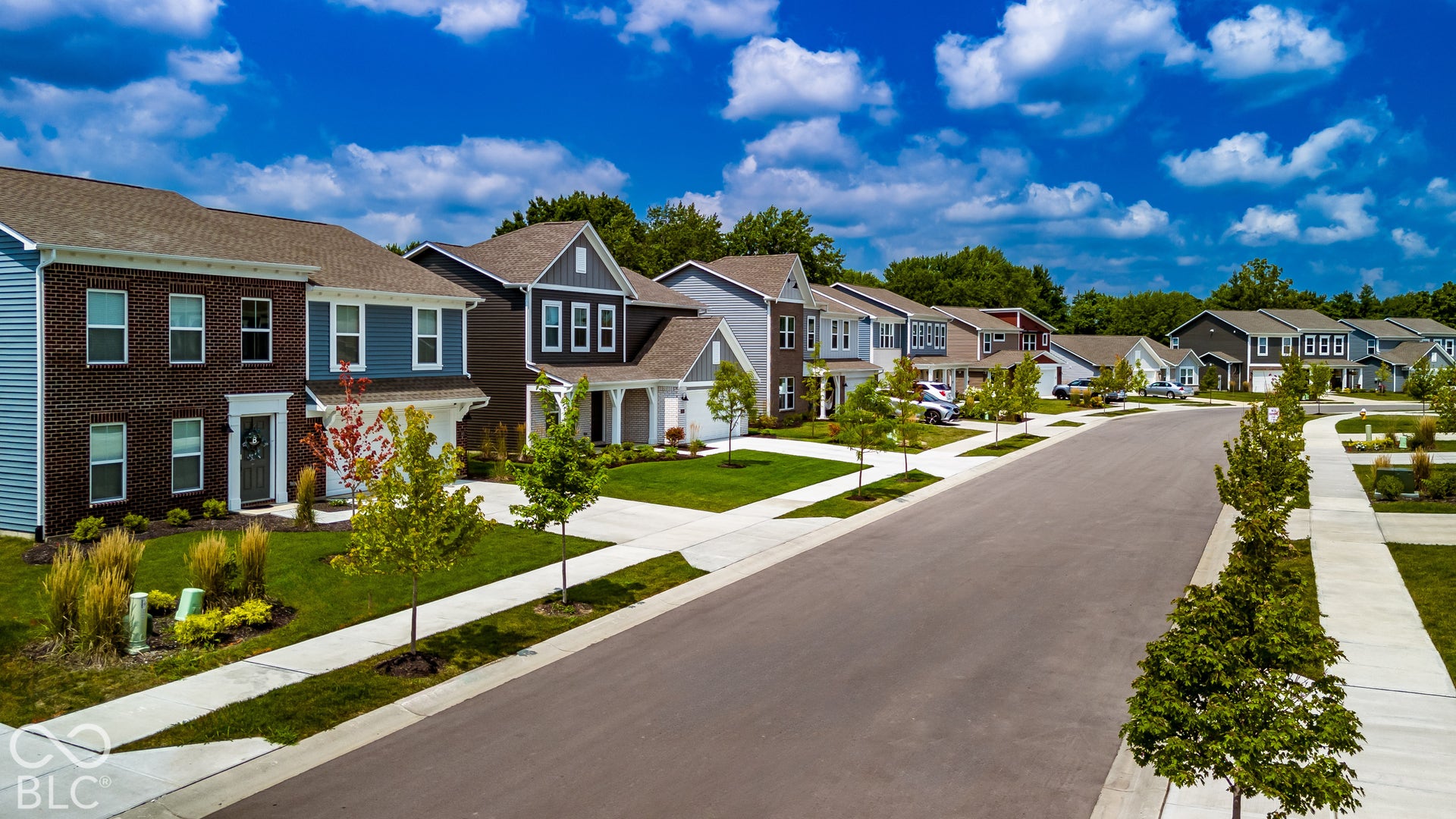 Suburban streetscape featuring multi-story homes with brick and siding exteriors, concrete driveways, paved sidewalks, and manicured front lawns