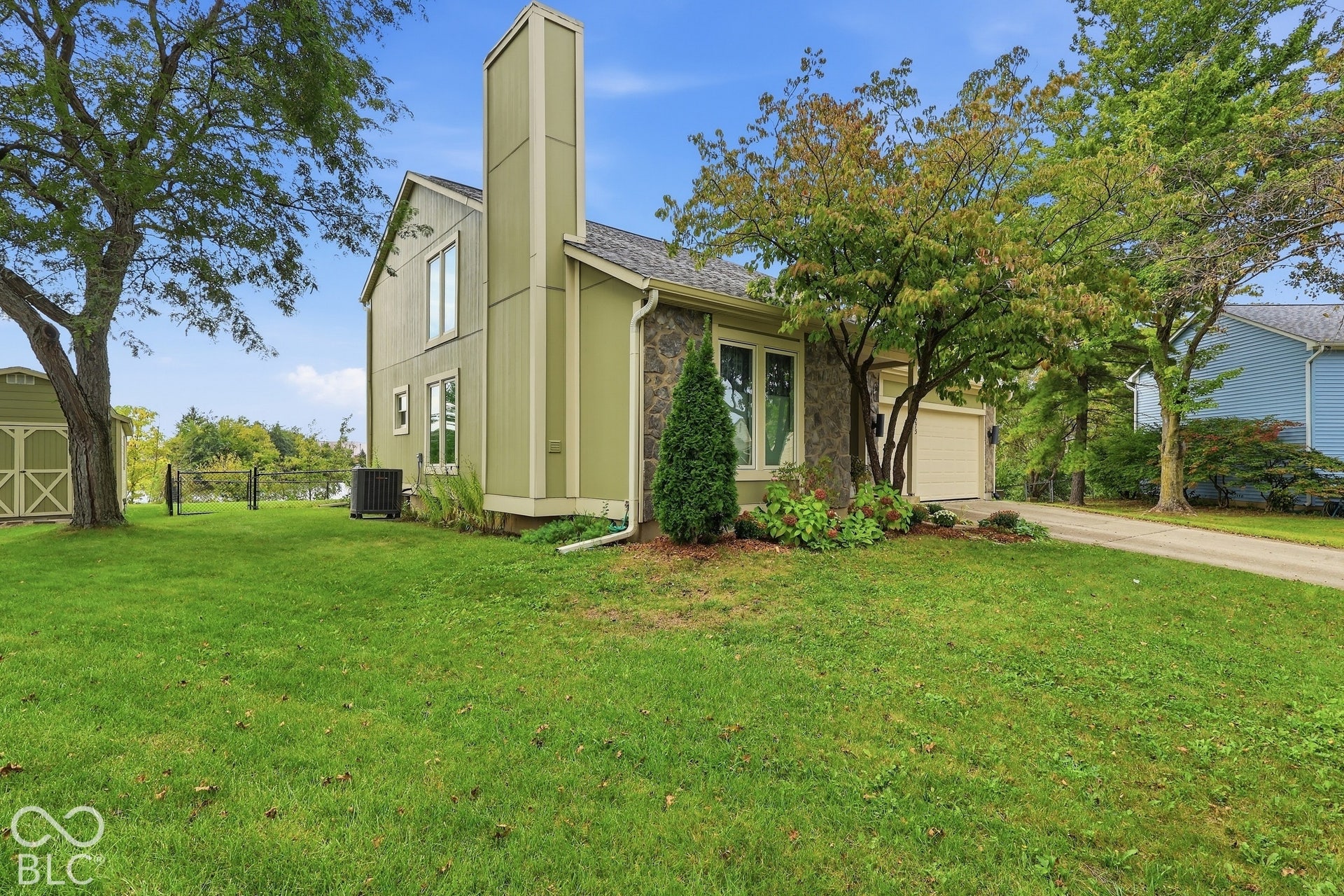 Exterior featuring stone siding accents, an attached two-car garage, an elongated chimney, and a detached storage structure on a grass-covered lot