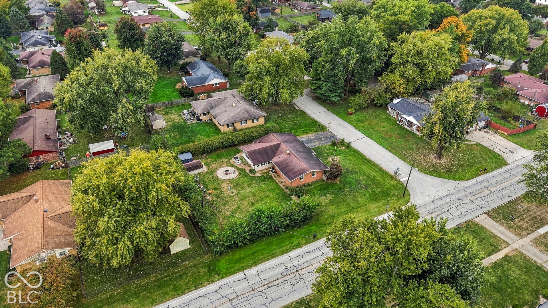 Aerial perspective featuring a brick exterior residence, a circular stone fire pit, mature trees, and an expansive lawn