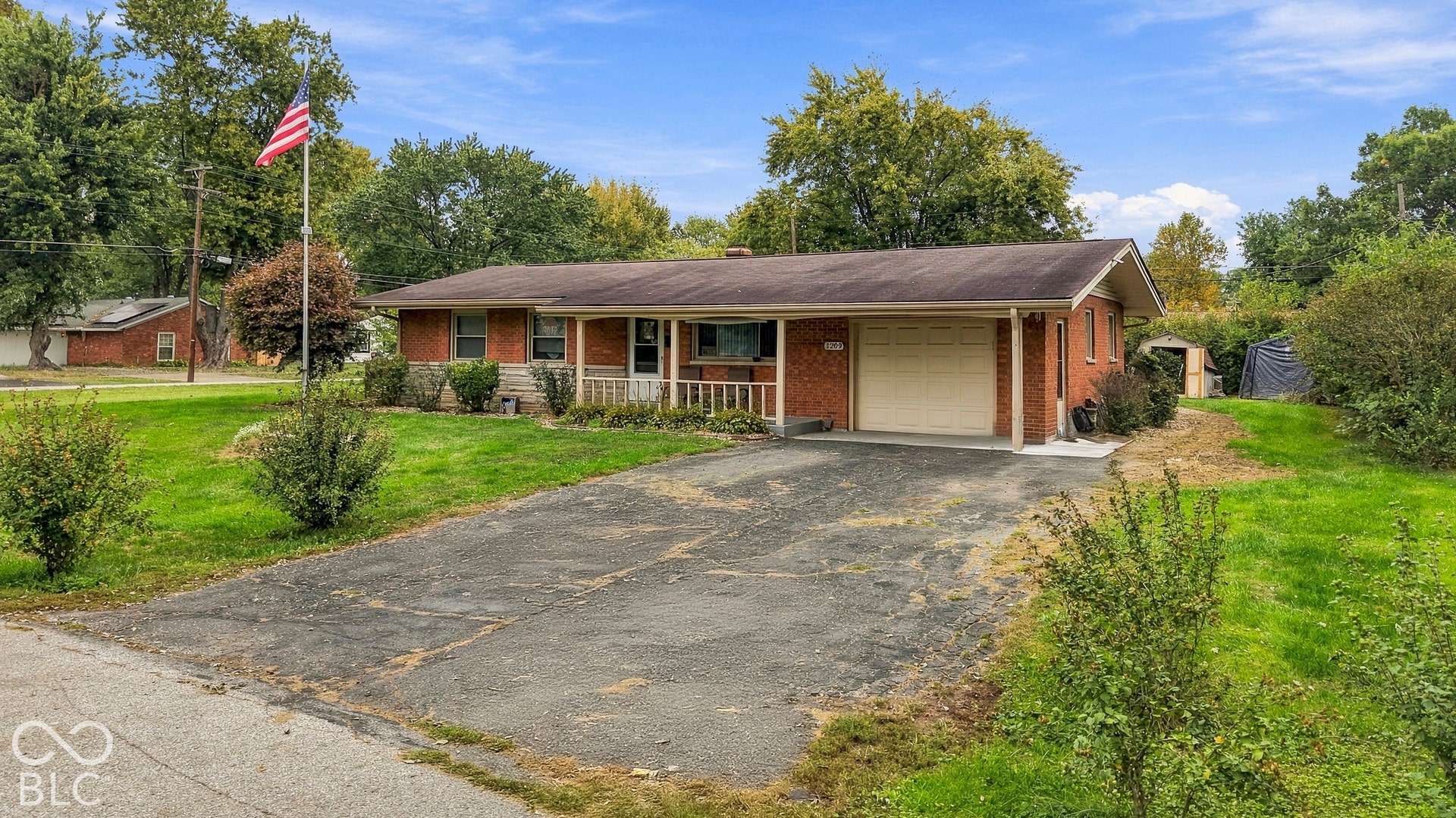 The exterior front of the ranch-style home featuring a covered front porch, an attached garage, and a concrete driveway on a landscaped lot. Flag pole in the center of the front yard.